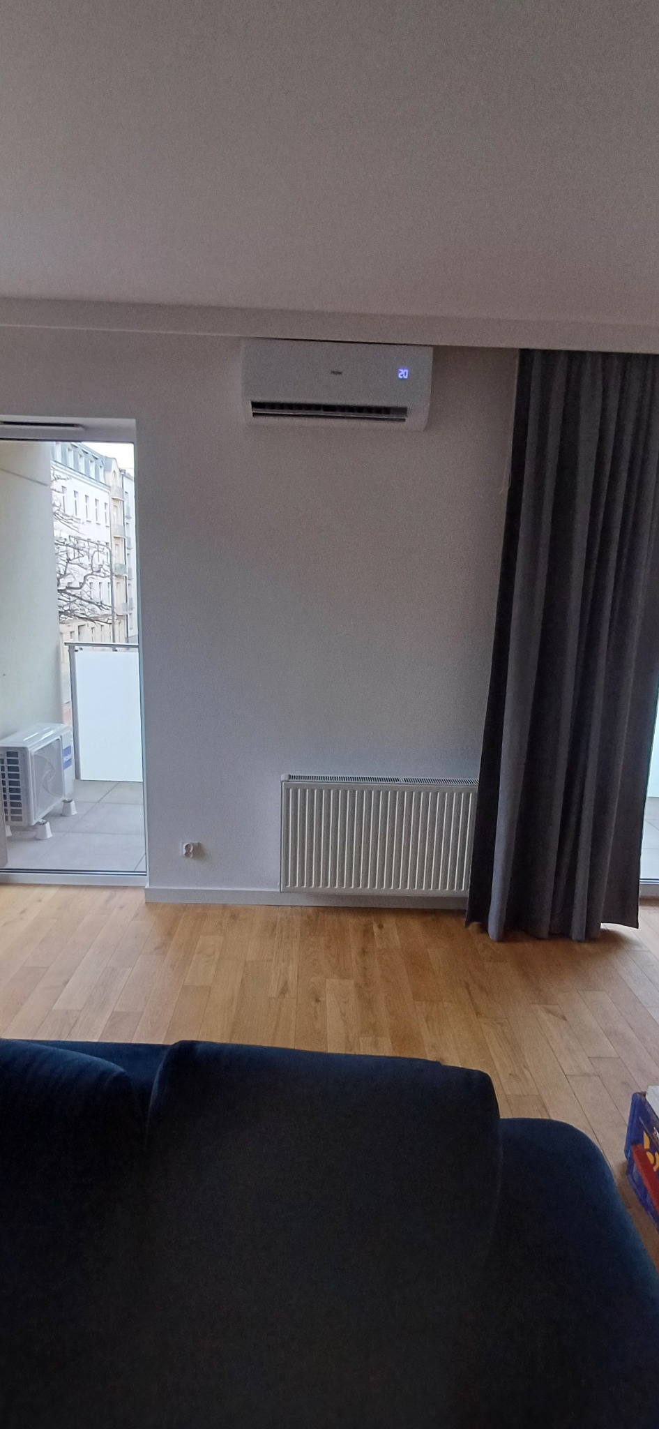 Biały klimatyzator z wyświetlaczem temperatury zamontowany na ścianie w pokoju z grzejnikiem i zasłoniętym oknem balkonowym.