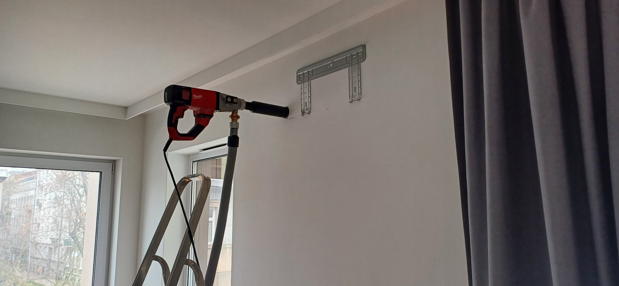 Czerwony młot udarowy Milwaukee z odkurzaczem wierci otwór w białej ścianie nad drabinką, obok okna i szarej zasłony, przygotowanie do montażu klimatyzacji.