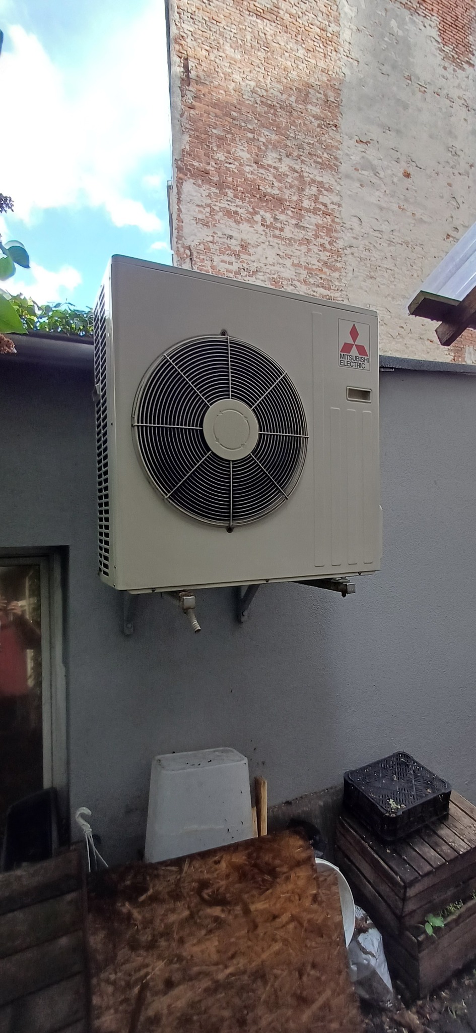 Biała jednostka zewnętrzna klimatyzacji Mitsubishi Electric, zamontowana na szarej ścianie budynku. Widoczne elementy instalacji i otoczenie.
