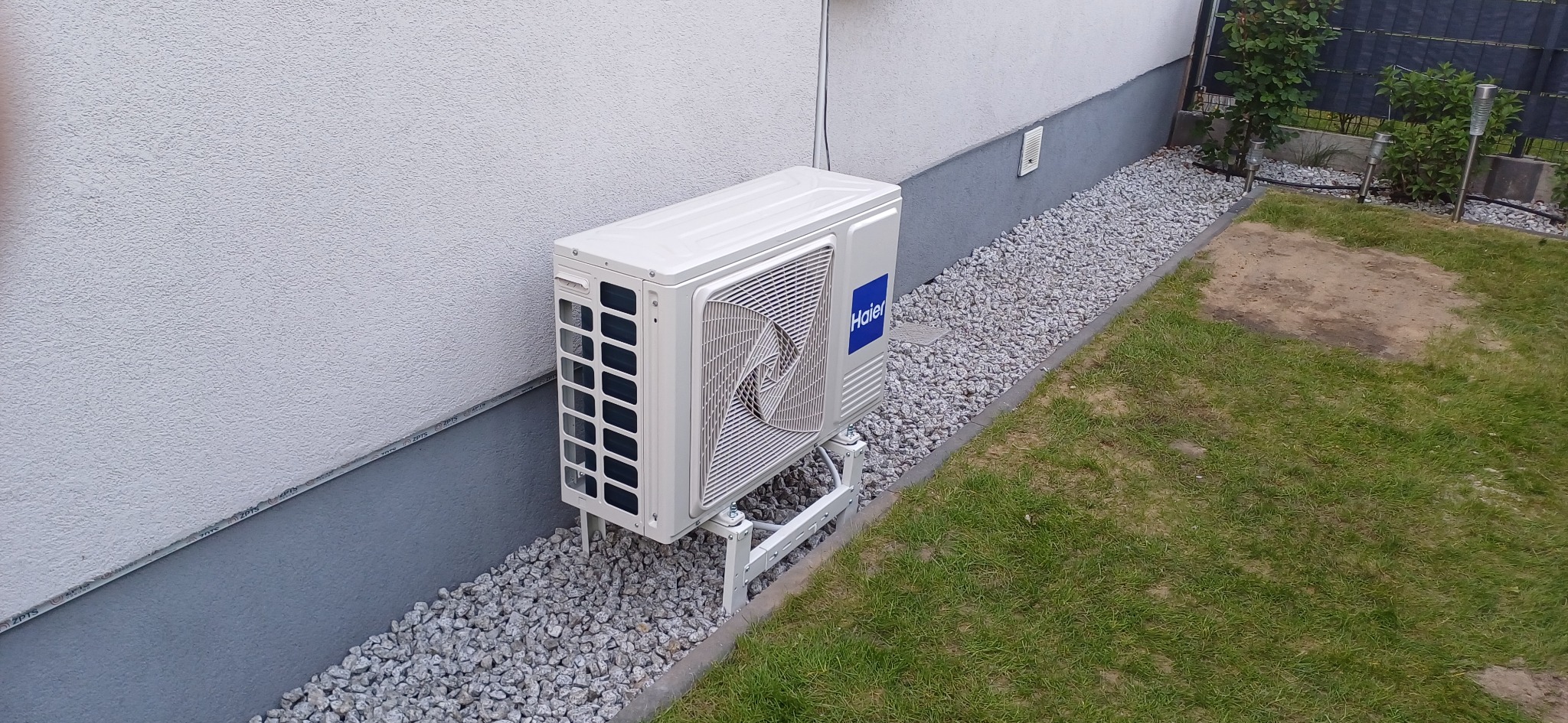Biały klimatyzator Haier zamontowany na zewnątrz budynku, na metalowym stelażu, z widocznym fragmentem trawnika i ozdobnymi kamieniami.