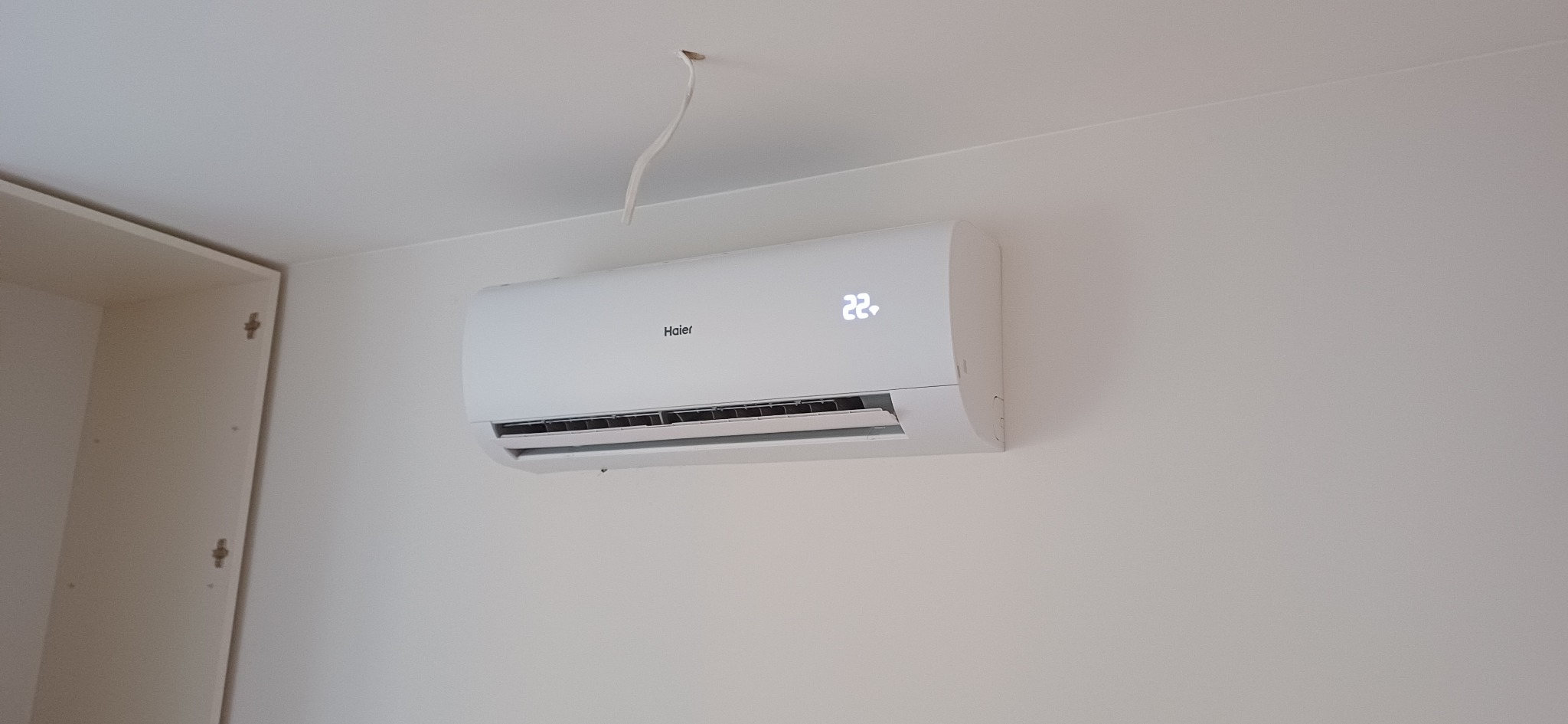 Biały klimatyzator Haier z widocznym wyświetlaczem temperatury 22°C, zamontowany na jasnej ścianie w pomieszczeniu, z fragmentem szafy po lewej stronie.