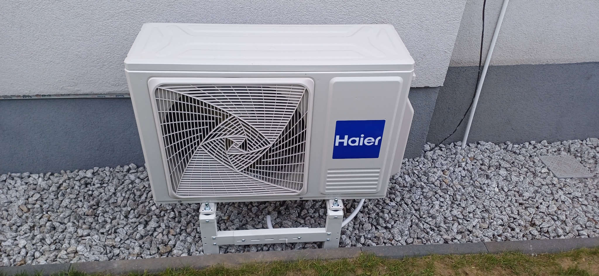 Biała jednostka zewnętrzna klimatyzacji Haier, zamontowana na elewacji budynku na niskim stelażu, otoczona kamieniami ozdobnymi.