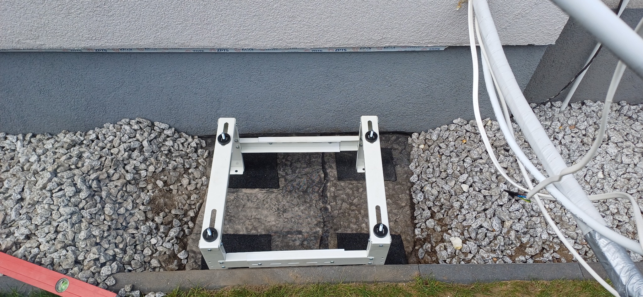 Biała rama montażowa klimatyzatora na podłożu z kamieni i czarnych płyt, przy szarej ścianie budynku. Widoczne przewody i poziomica.