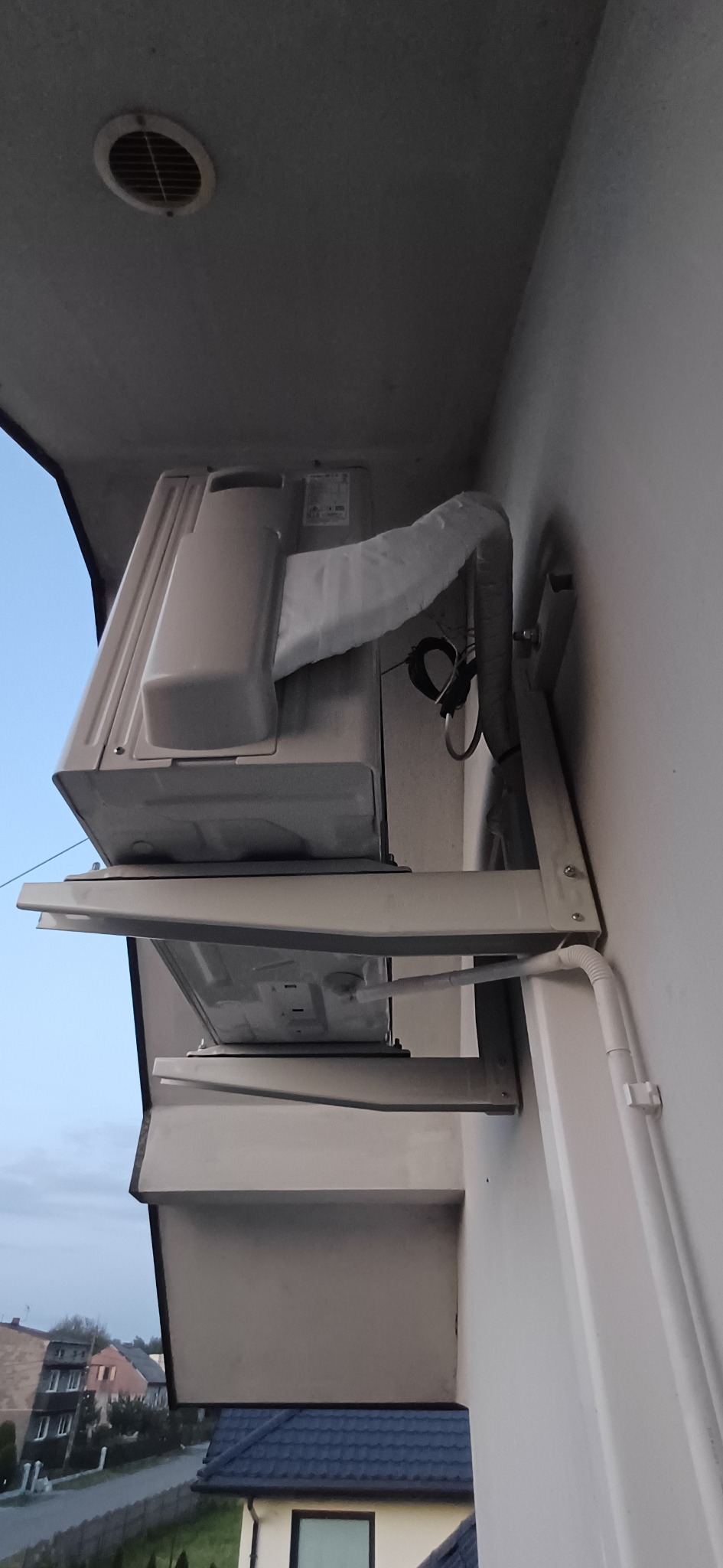 Zamontowana jednostka zewnętrzna klimatyzacji na wspornikach przy elewacji domu. Widoczne przewody i rury odprowadzające skropliny. Ujęcie z dołu, widoczne fragmenty dachu.