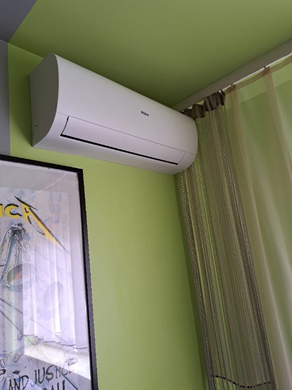 Biała klimatyzacja Haier na zielonej ścianie, obok firanka i plakat. Widok z dołu, oświetlenie dzienne. Nowoczesny design.