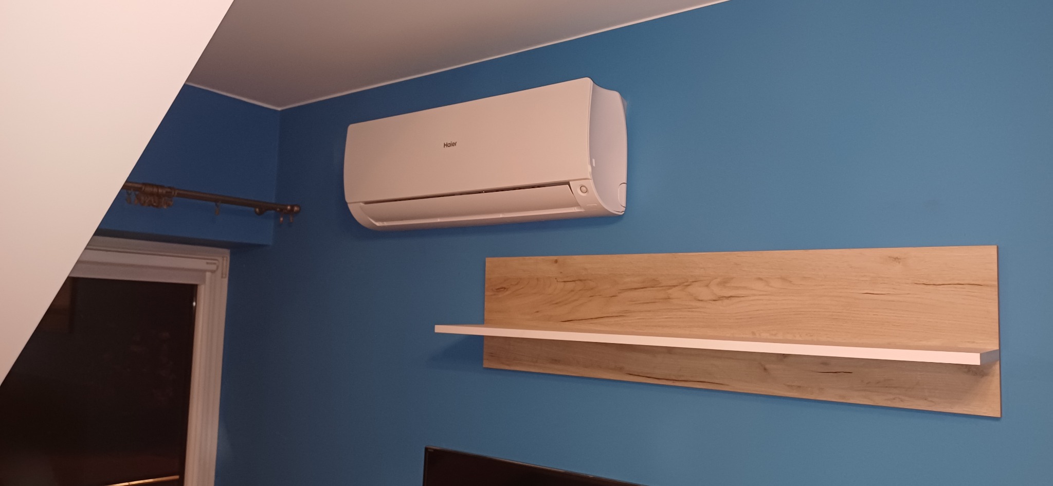 Biały klimatyzator Haier zamontowany na niebieskiej ścianie nad drewnianą półką w pokoju. Widoczny fragment karnisza i telewizora.