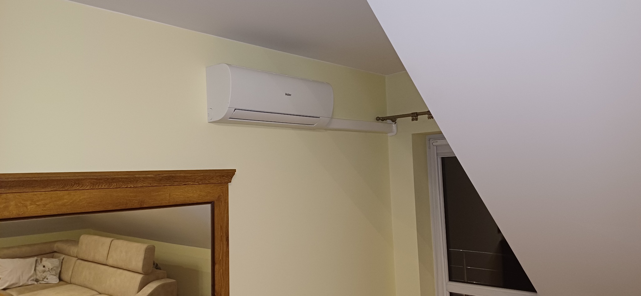 Biały klimatyzator Haier zamontowany na żółtej ścianie w pokoju z beżową sofą i oknem. Widoczny fragment ramy lustra.