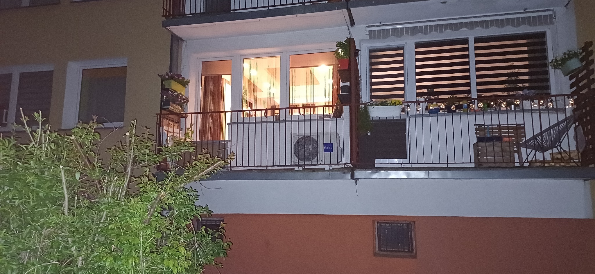 Balkon z klimatyzatorem Haier, kwiatami w donicach i meblami. Widok z dołu, oświetlenie wieczorne.