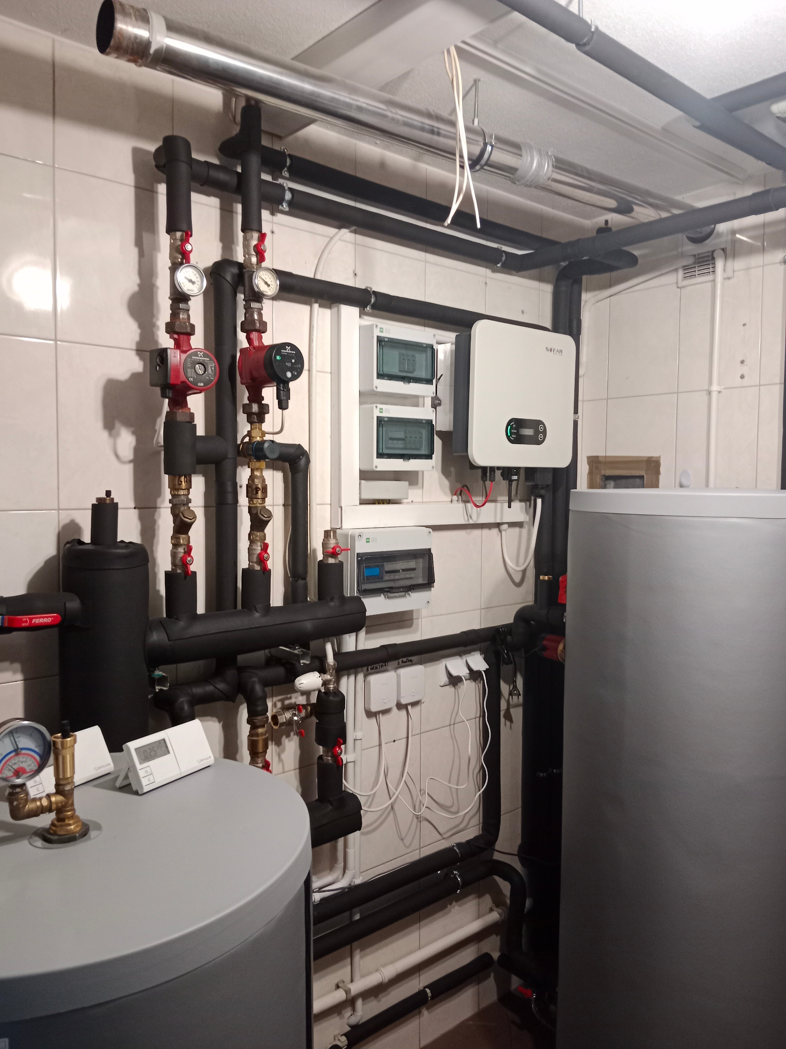 Wnętrze kotłowni z zainstalowanym zasobnikiem ciepłej wody, pompami obiegowymi, orurowaniem w izolacji, rozdzielnicami elektrycznymi i inwerterem fotowoltaicznym na kafelkowanej ścianie.