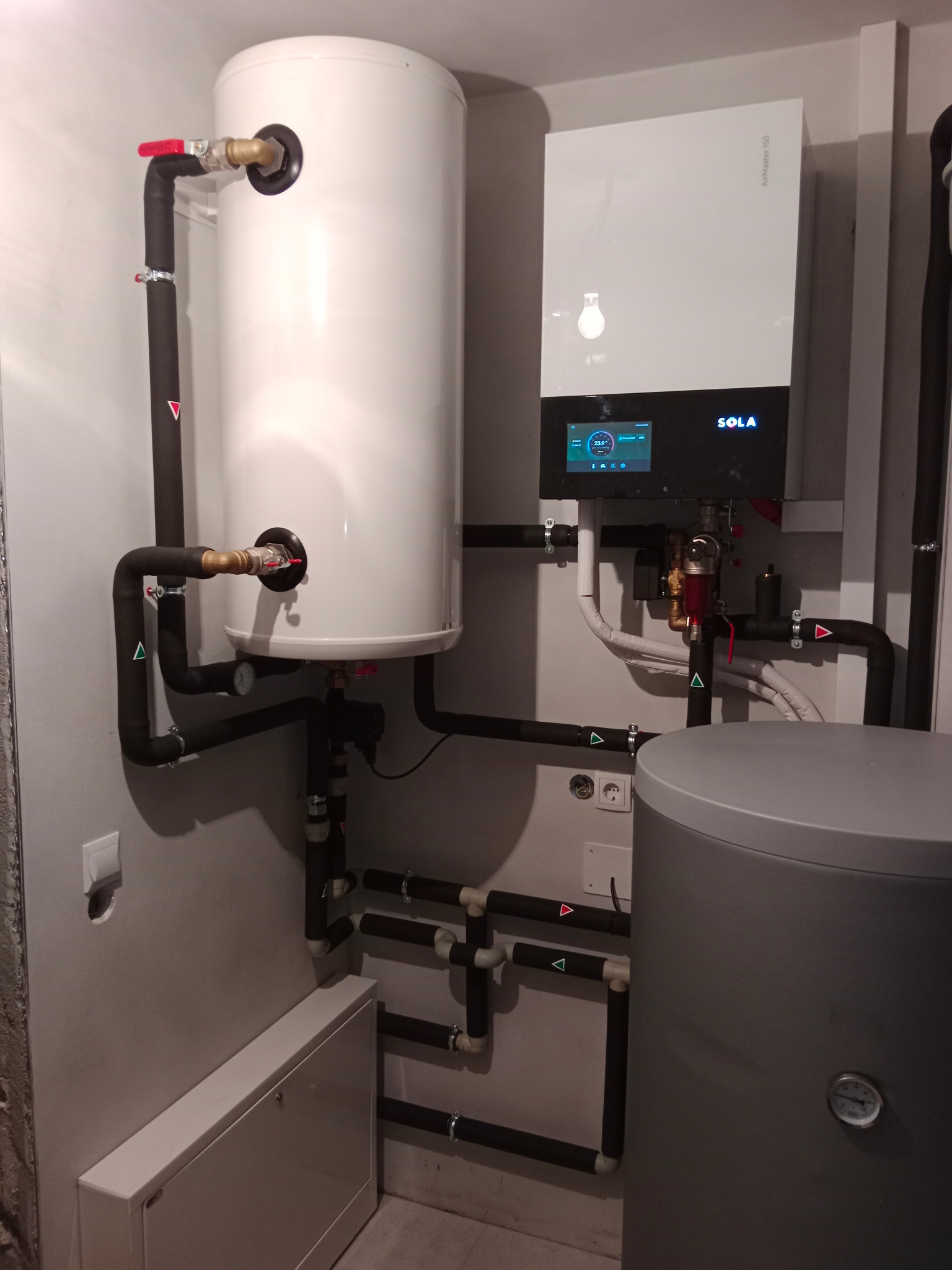Biały zasobnik ciepłej wody użytkowej i wiszący kocioł grzewczy Sola AirMaster ISO z wyświetlaczem, instalacja rur w izolacji z oznaczeniami, w tle szary zbiornik akumulacyjny.