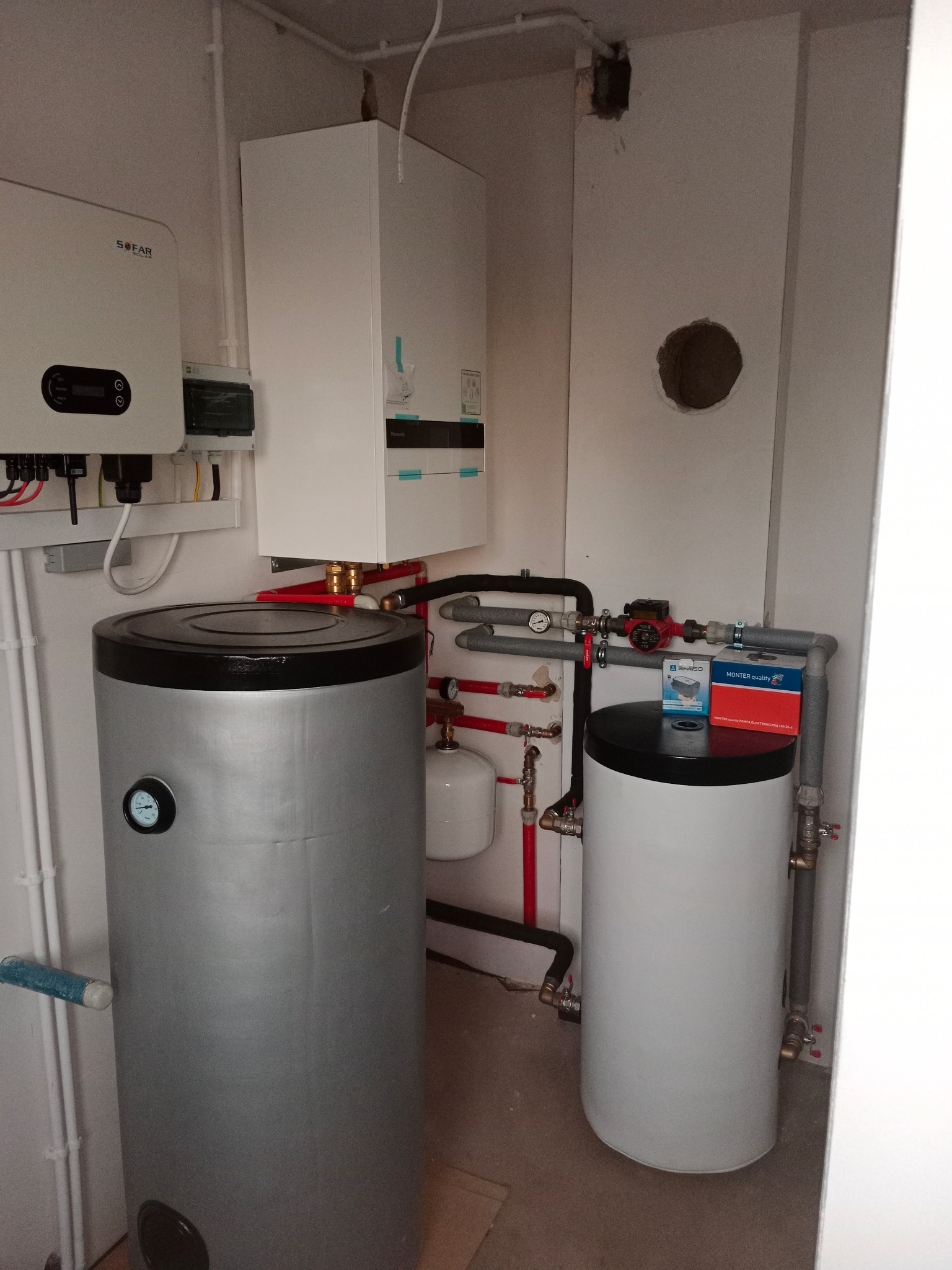 W kotłowni widoczny srebrny bojler z czarnym wiekiem, obok biały bojler, instalacja z rur w kolorze czerwonym i szarym, na ścianie wisi inwerter SOFAR i biały piec, ściana z dziurą na instalacje.