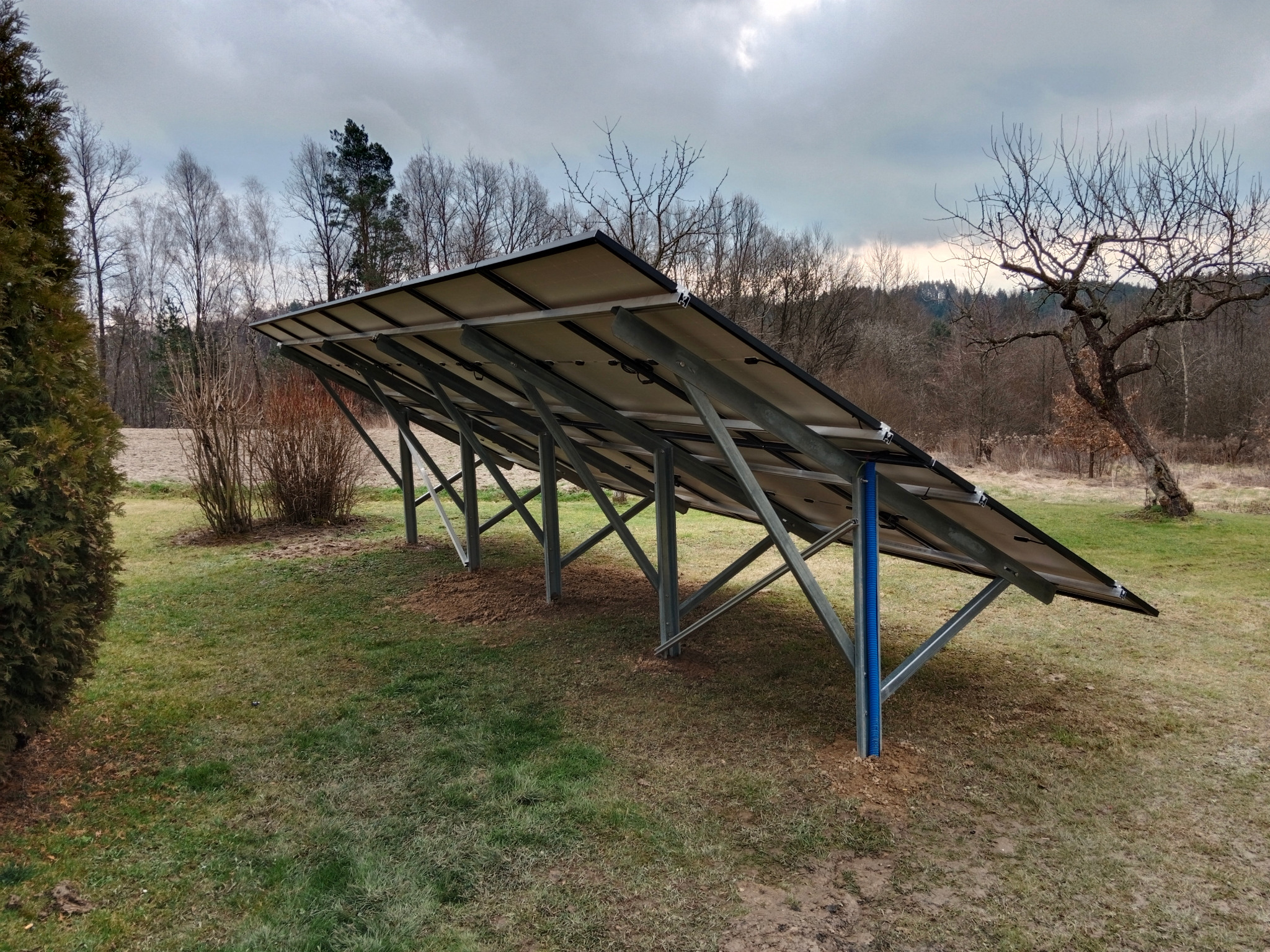 Rząd paneli słonecznych zamontowanych na metalowej konstrukcji, widok od spodu, z widocznymi niebieskimi rurami osłonowymi na tle trawnika i drzew bez liści.