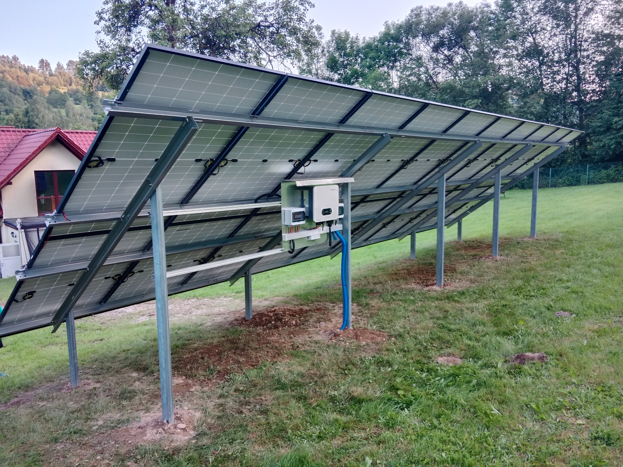 Widok od tyłu na panele słoneczne zamontowane na metalowej konstrukcji na trawiastym terenie, z widocznym falownikiem i okablowaniem, w tle dom z czerwonym dachem i drzewa.