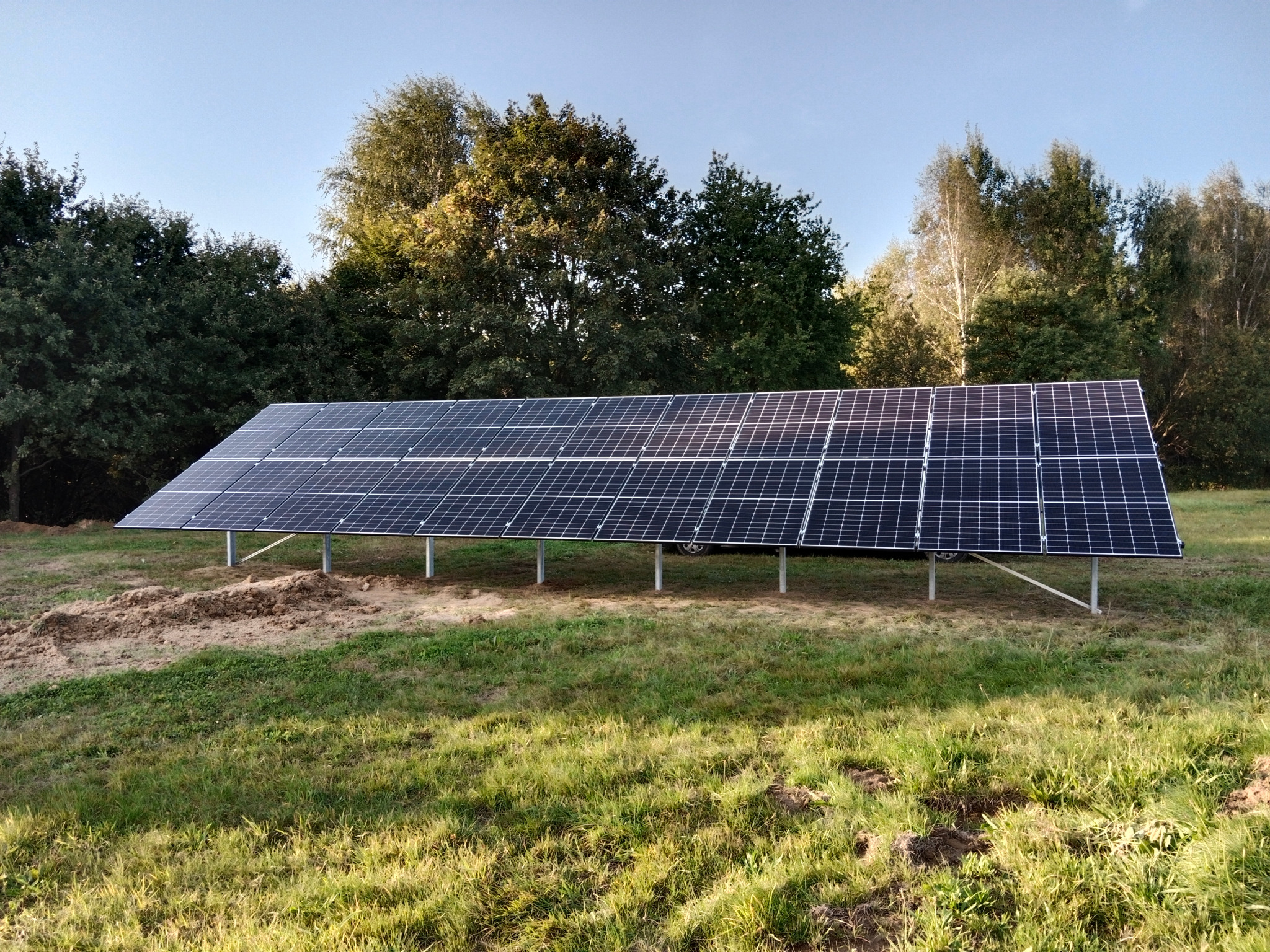 Instalacja fotowoltaiczna na trawiastym terenie w Rzepienniku Strzyżewskim, panele zamontowane na metalowej konstrukcji, w tle drzewa i błękitne niebo.