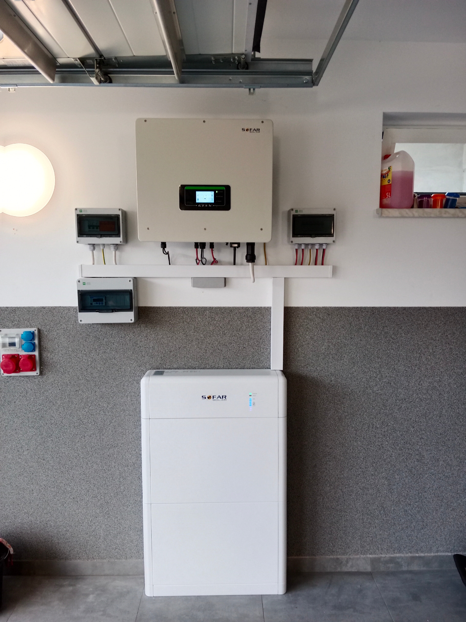 Biały inwerter i akumulator SOFAR zamontowane na ścianie w garażu, widoczne skrzynki elektryczne, przewody i rury instalacyjne.