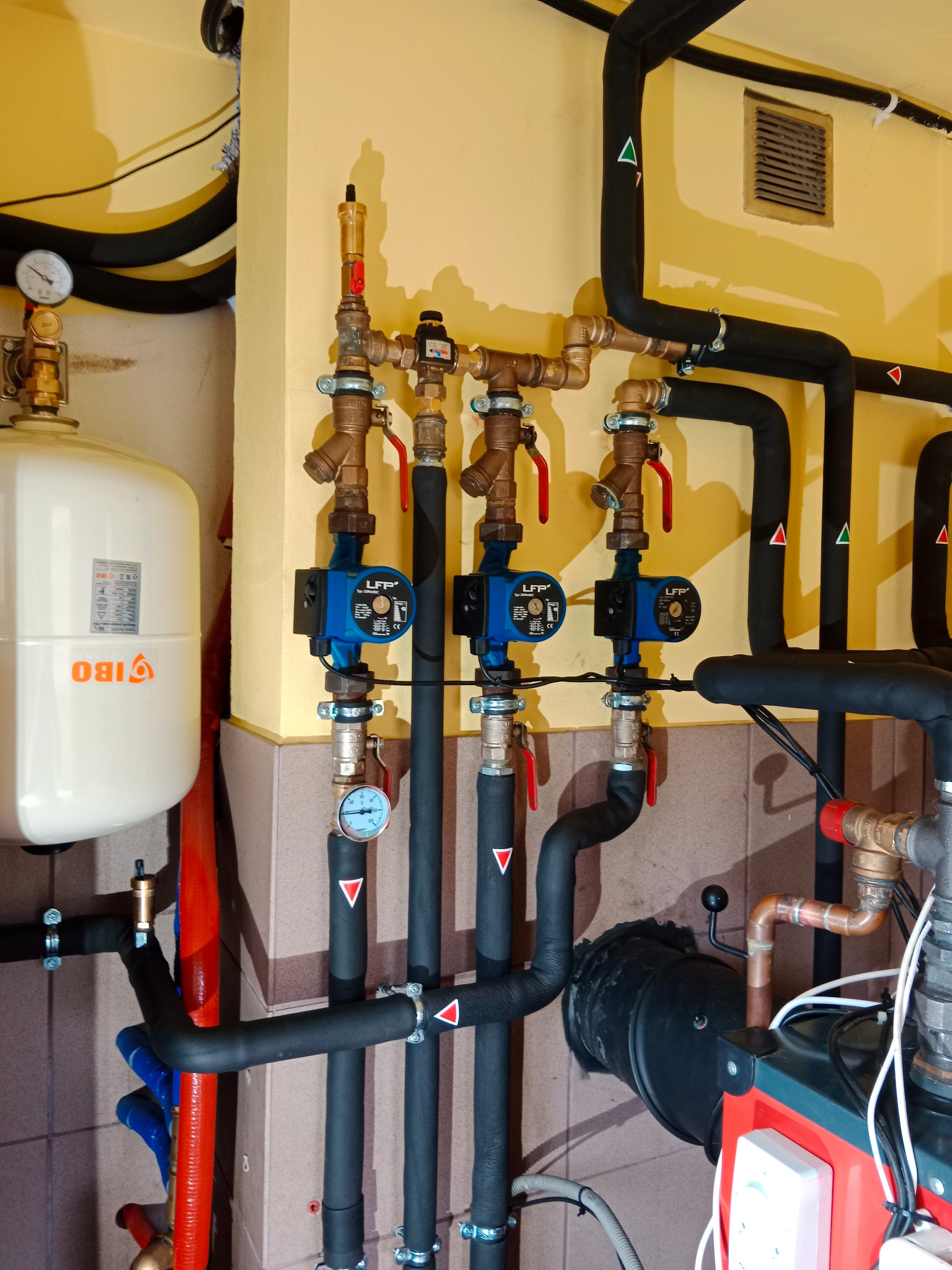 Węzeł cieplny z trzema pompami obiegowymi LFP, manometrami, zaworami, rurami w czarnej izolacji z oznaczeniami kolorystycznymi, zbiornikiem przeponowym IBO oraz elementami kotła grzewczego...