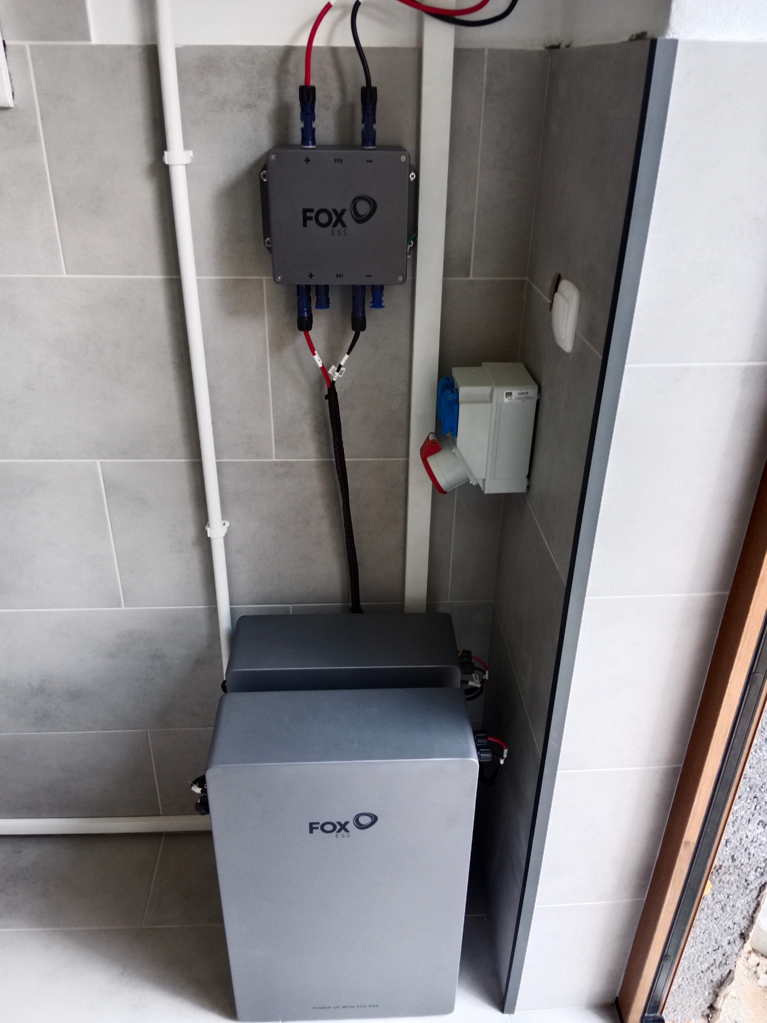 Dwa szare akumulatory Fox ESS ustawione jeden na drugim, podłączone do szarej skrzynki z kablami na ścianie z szarych płytek, widoczny biały przewód i gniazdo elektryczne.