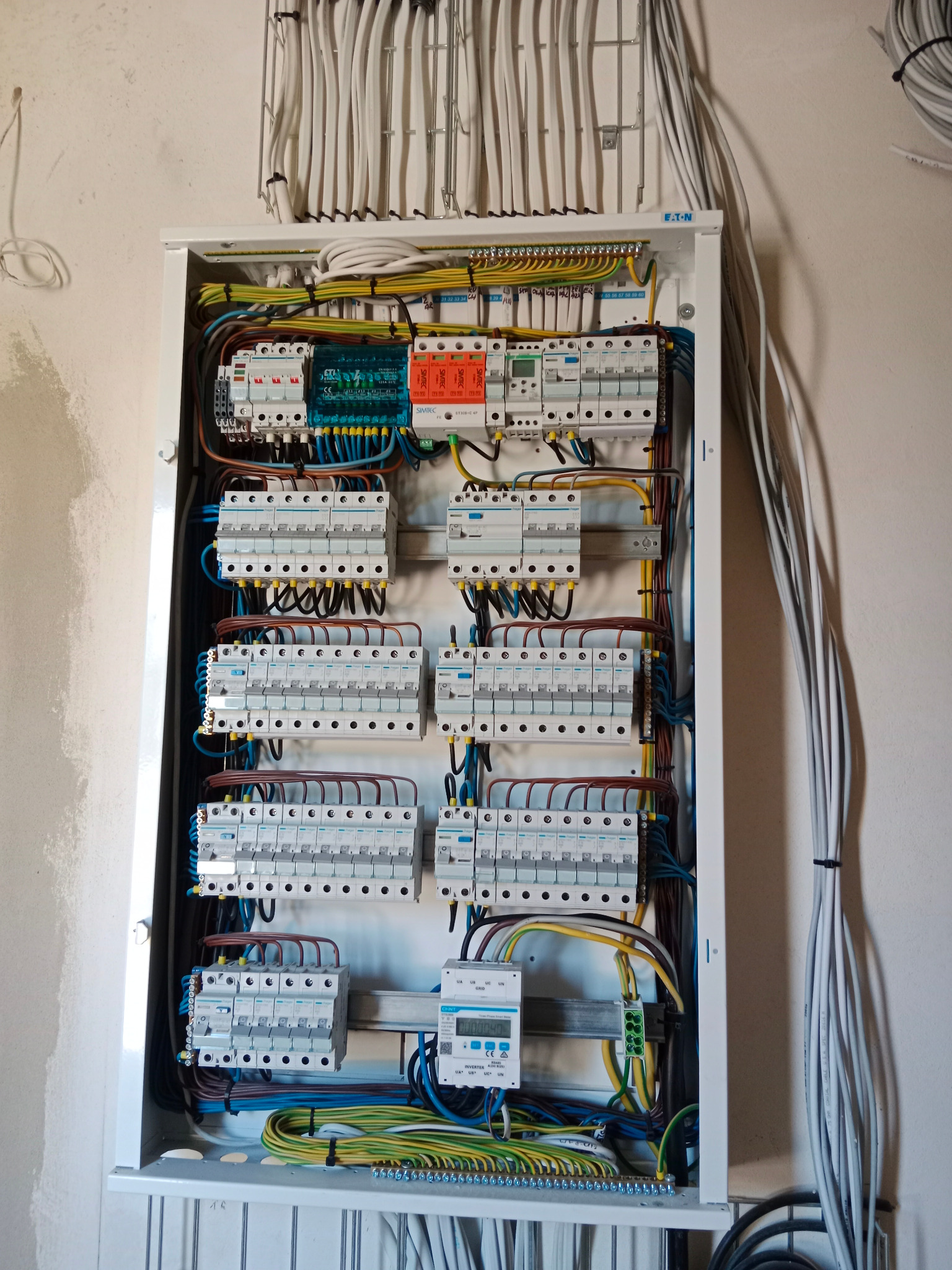 Otwarta skrzynka elektryczna z licznymi bezpiecznikami, przewodami w różnych kolorach i miernikiem, zamontowana na ścianie w surowym stanie.
