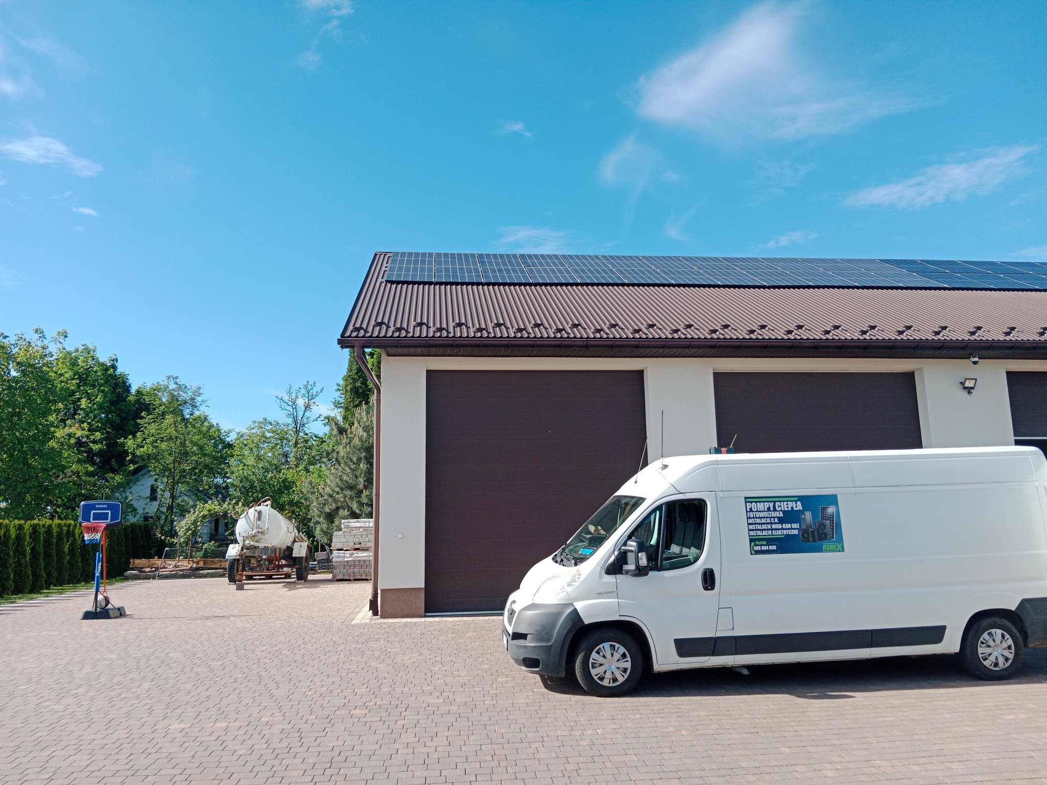 Biały samochód dostawczy z reklamą firmy instalacyjnej zaparkowany przed budynkiem z panelami słonecznymi na dachu, na tle błękitnego nieba. W tle widoczny kosz do koszykówki i beczkowóz.