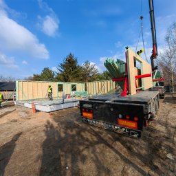 REALGREEN.HOUSE SPÓŁKA Z OGRANICZONĄ ODPOWIEDZIALNOŚCIĄ - Montaż drewnianej konstrukcji domu z użyciem dźwigu na fundamencie, widoczni pracownicy w odblaskowych kamizelkach i kaskach.