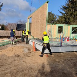 REALGREEN.HOUSE SPÓŁKA Z OGRANICZONĄ ODPOWIEDZIALNOŚCIĄ - Montaż ściany szkieletowej domu jednorodzinnego z pomocą dźwigu; pracownicy w żółtych kaskach i kamizelkach odblaskowych na placu budowy.
