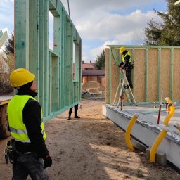 REALGREEN.HOUSE SPÓŁKA Z OGRANICZONĄ ODPOWIEDZIALNOŚCIĄ - Montaż ściany z drewnianej konstrukcji szkieletowej na fundamencie; widoczni pracownicy w kaskach i kamizelkach odblaskowych.