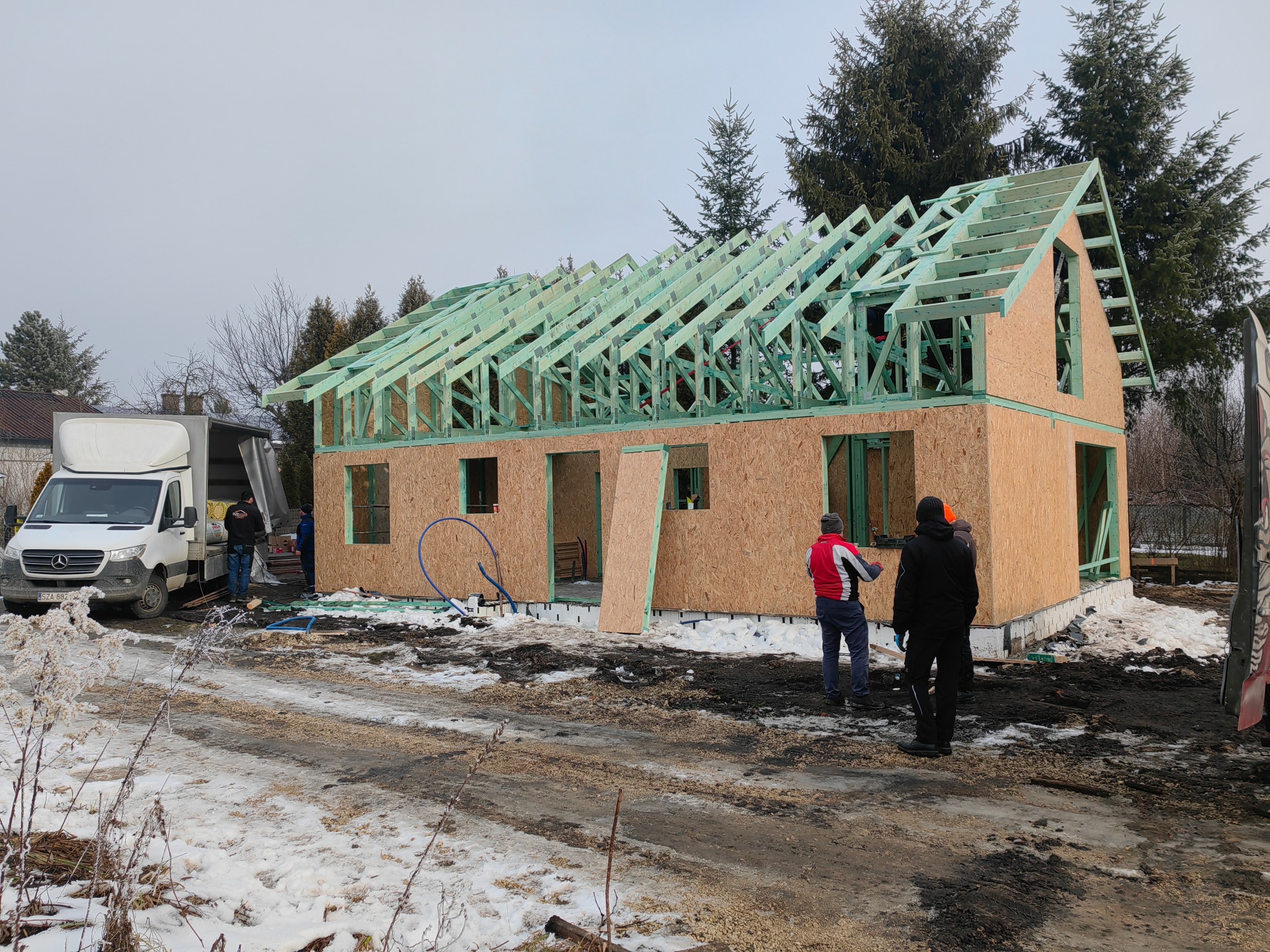 Wznoszenie dwukondygnacyjnego budynku w technologii szkieletowej: widoczna konstrukcja ścian z płyty OSB i zielona więźba dachowa, ekipa budowlana przy białym samochodzie dostawczym, zimowa...