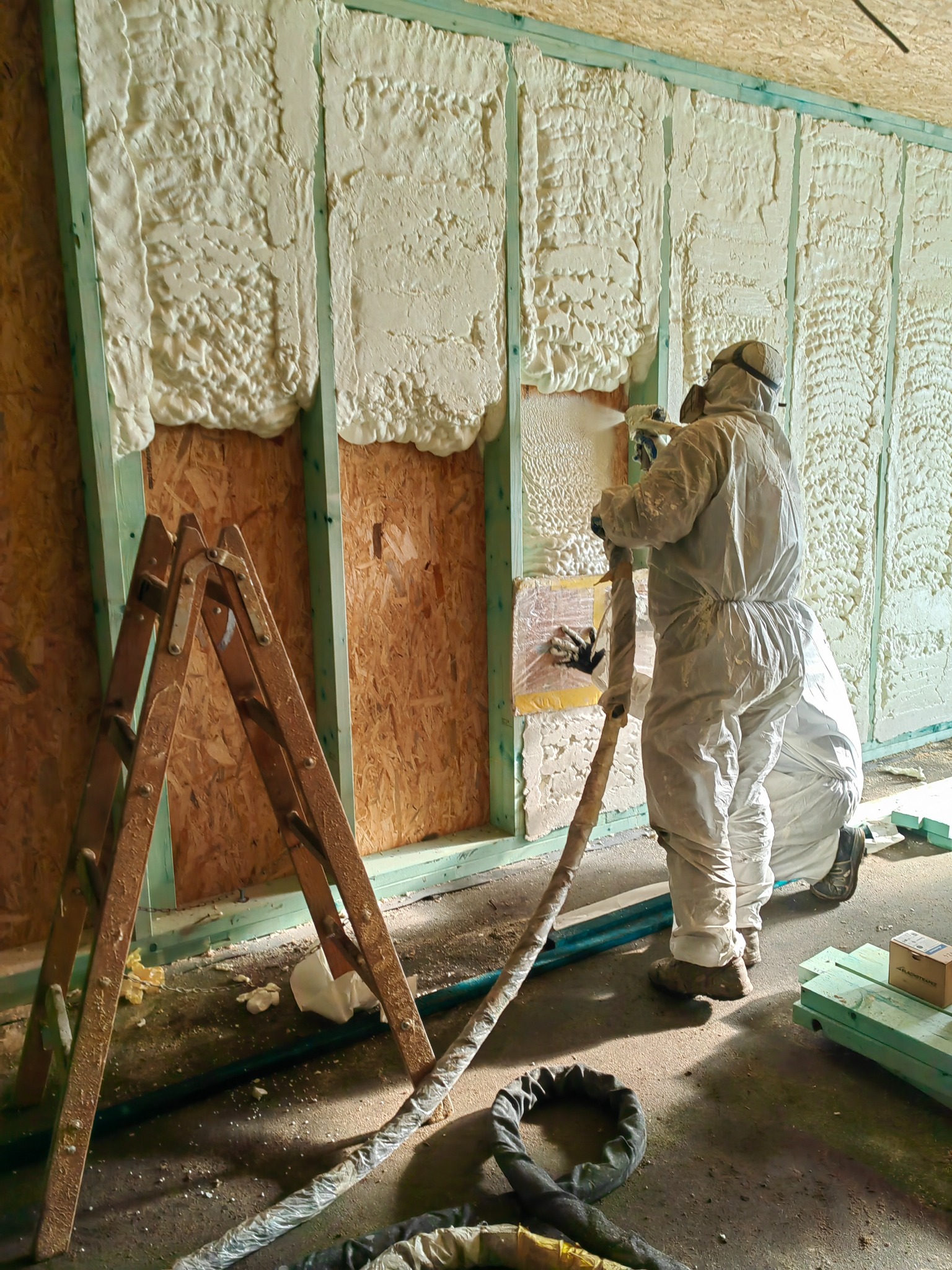 Pracownik w kombinezonie ochronnym natryskuje pianę izolacyjną na ścianę szkieletową z drewnianych belek, widoczna drabina i przewody.