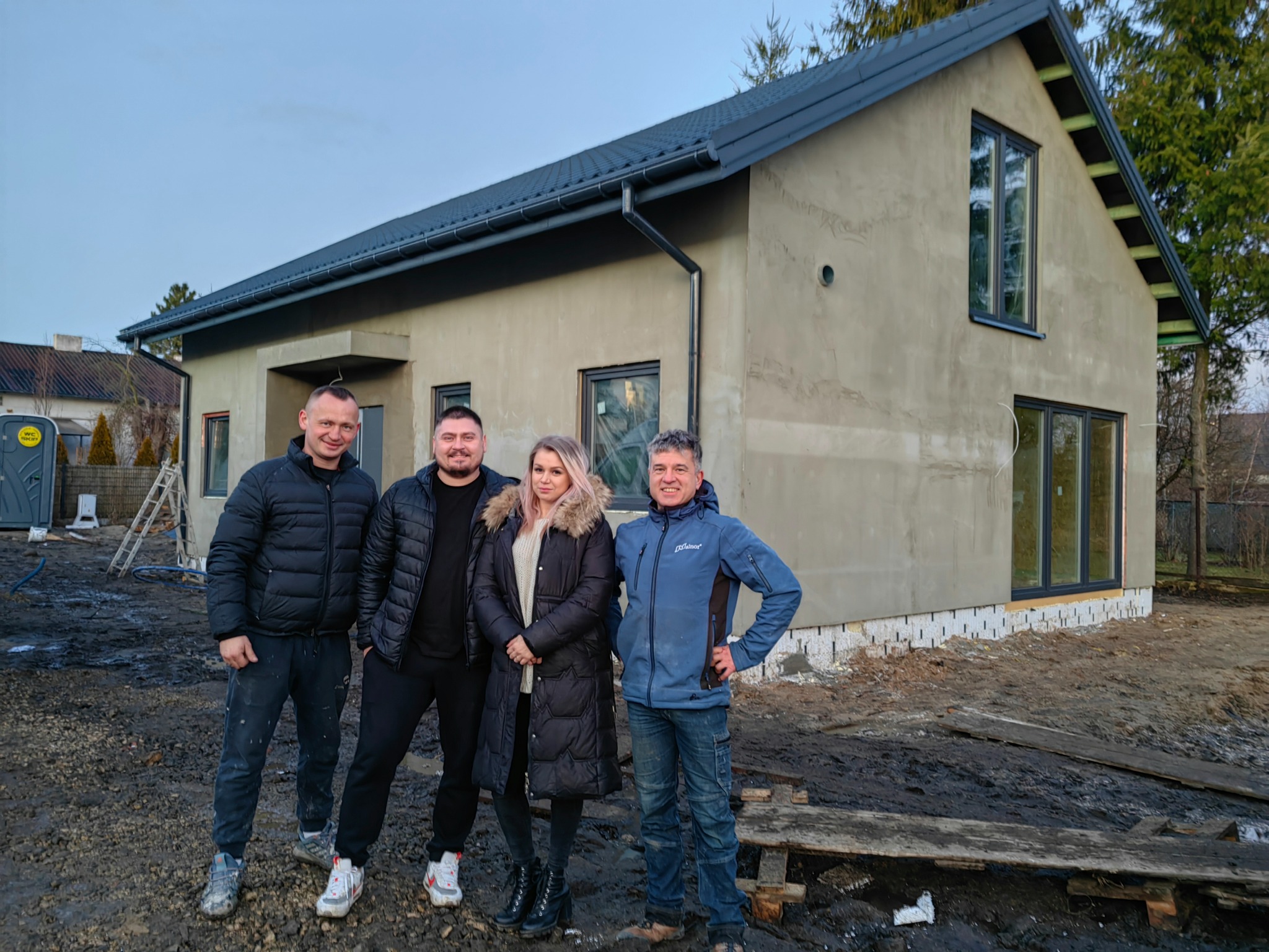 Ekipa budowlana pozuje przed nowoczesnym domem szkieletowym w trakcie wykończenia, widoczne szare ściany zewnętrzne i czarne okna.