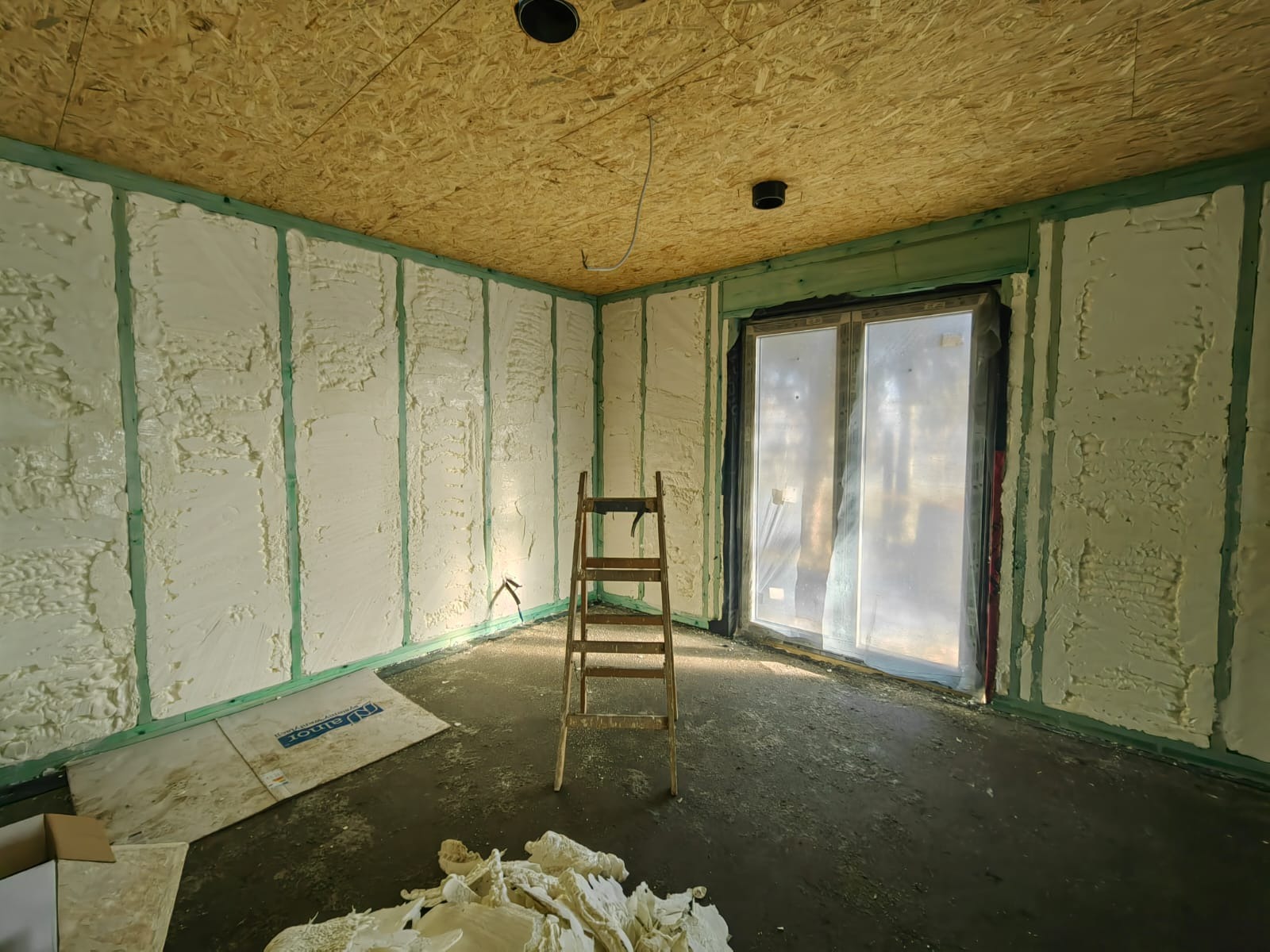 Wnętrze w trakcie izolacji pianą PUR w domu w konstrukcji szkieletowej, widoczne drewniane belki, drabina i folia ochronna na oknie.