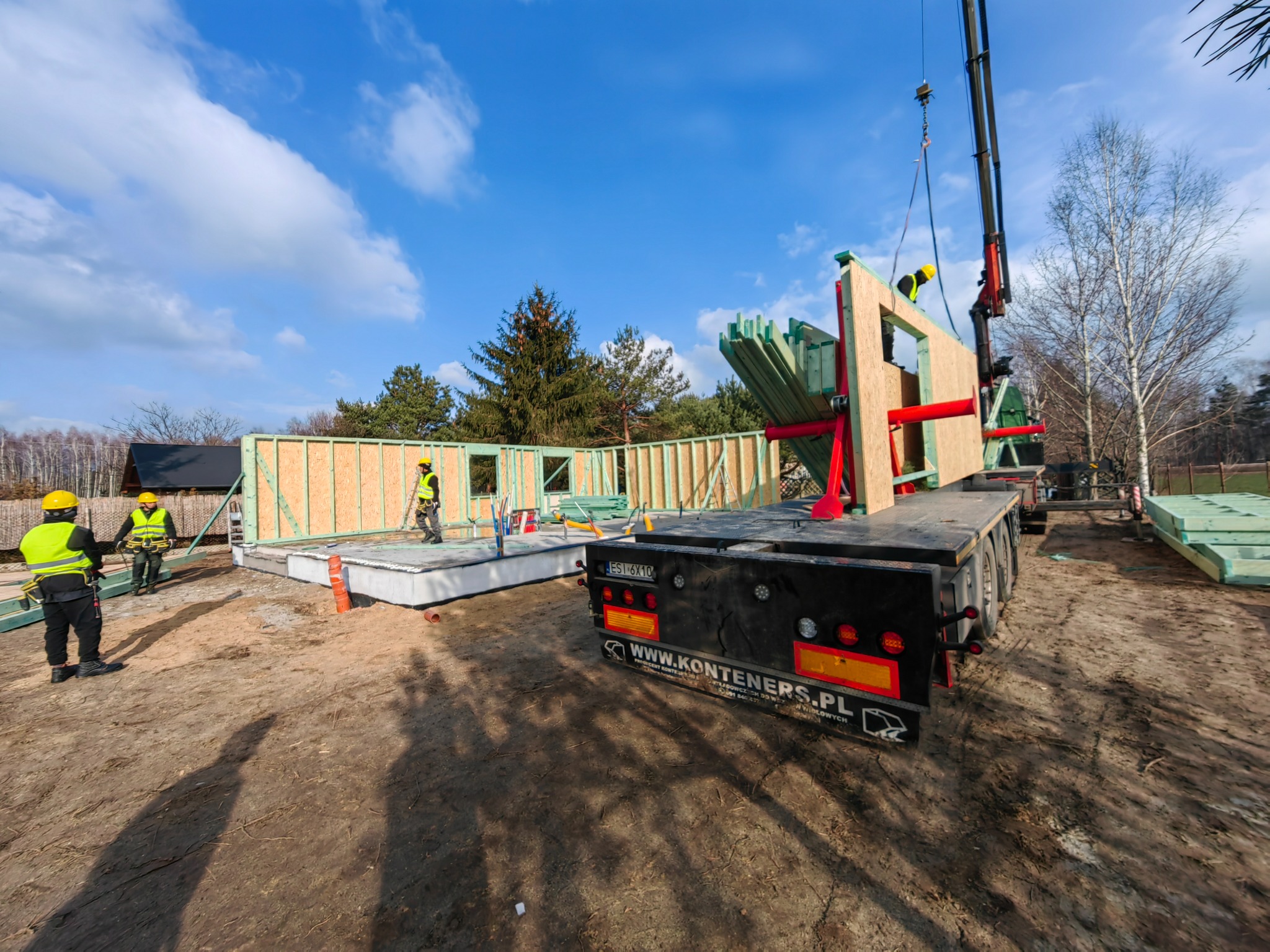 Montaż drewnianej konstrukcji domu z użyciem dźwigu na fundamencie, widoczni pracownicy w odblaskowych kamizelkach i kaskach.