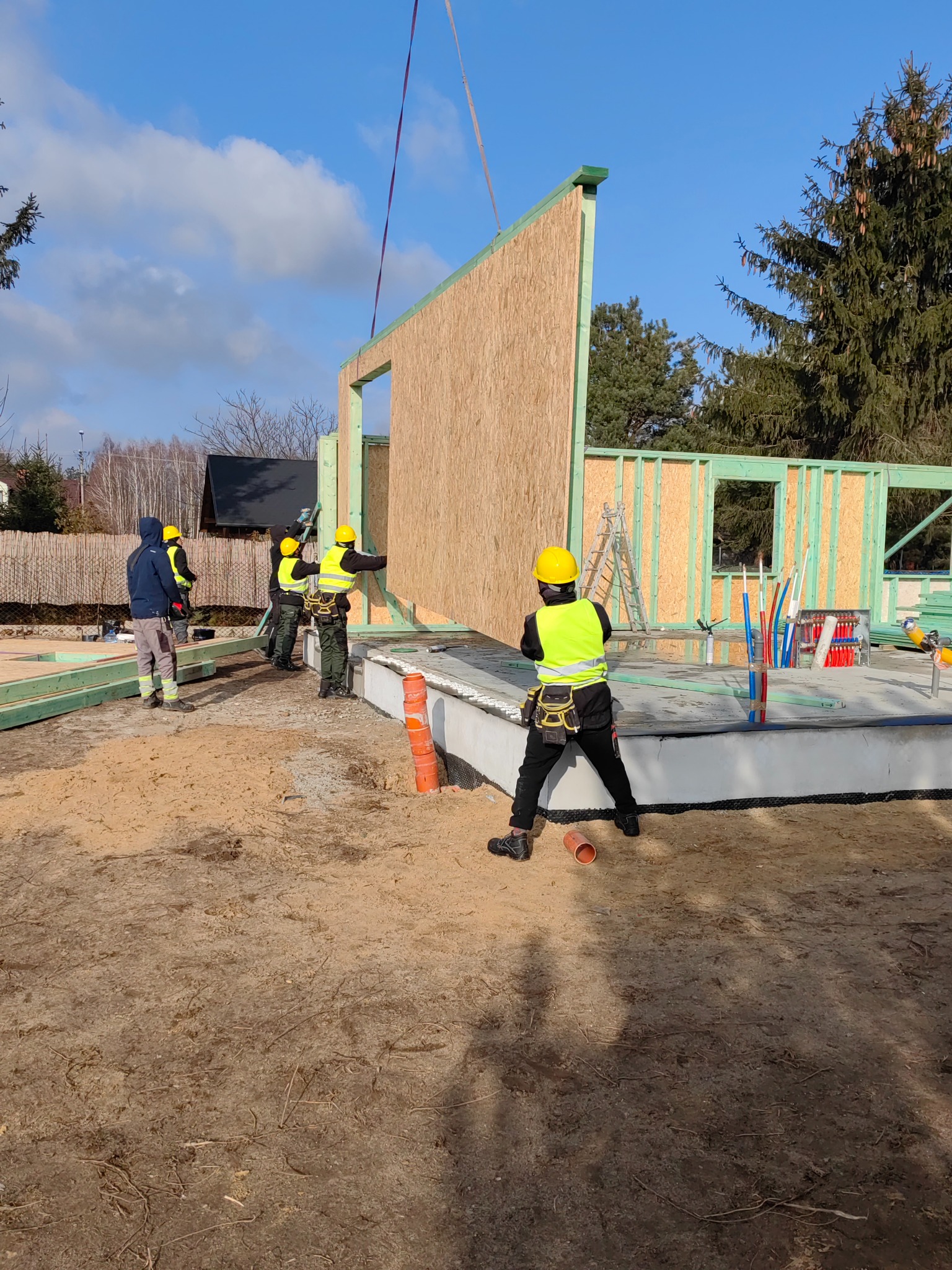 Montaż ściany szkieletowej domu jednorodzinnego z pomocą dźwigu; pracownicy w żółtych kaskach i kamizelkach odblaskowych na placu budowy.