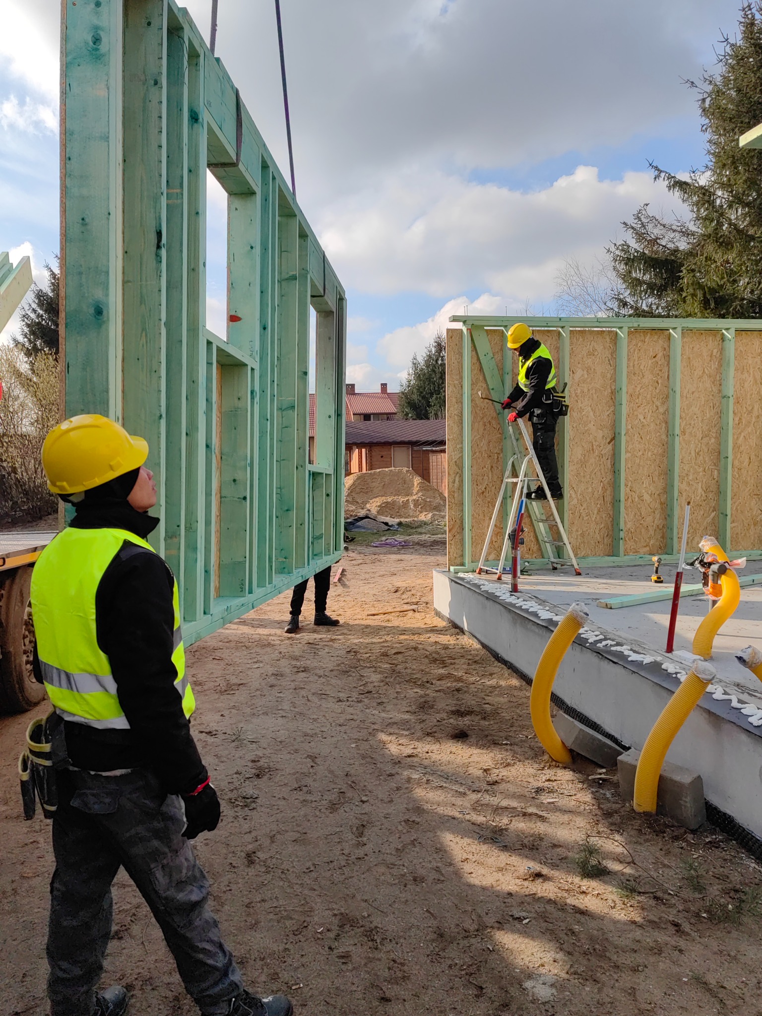 Montaż ściany z drewnianej konstrukcji szkieletowej na fundamencie; widoczni pracownicy w kaskach i kamizelkach odblaskowych.