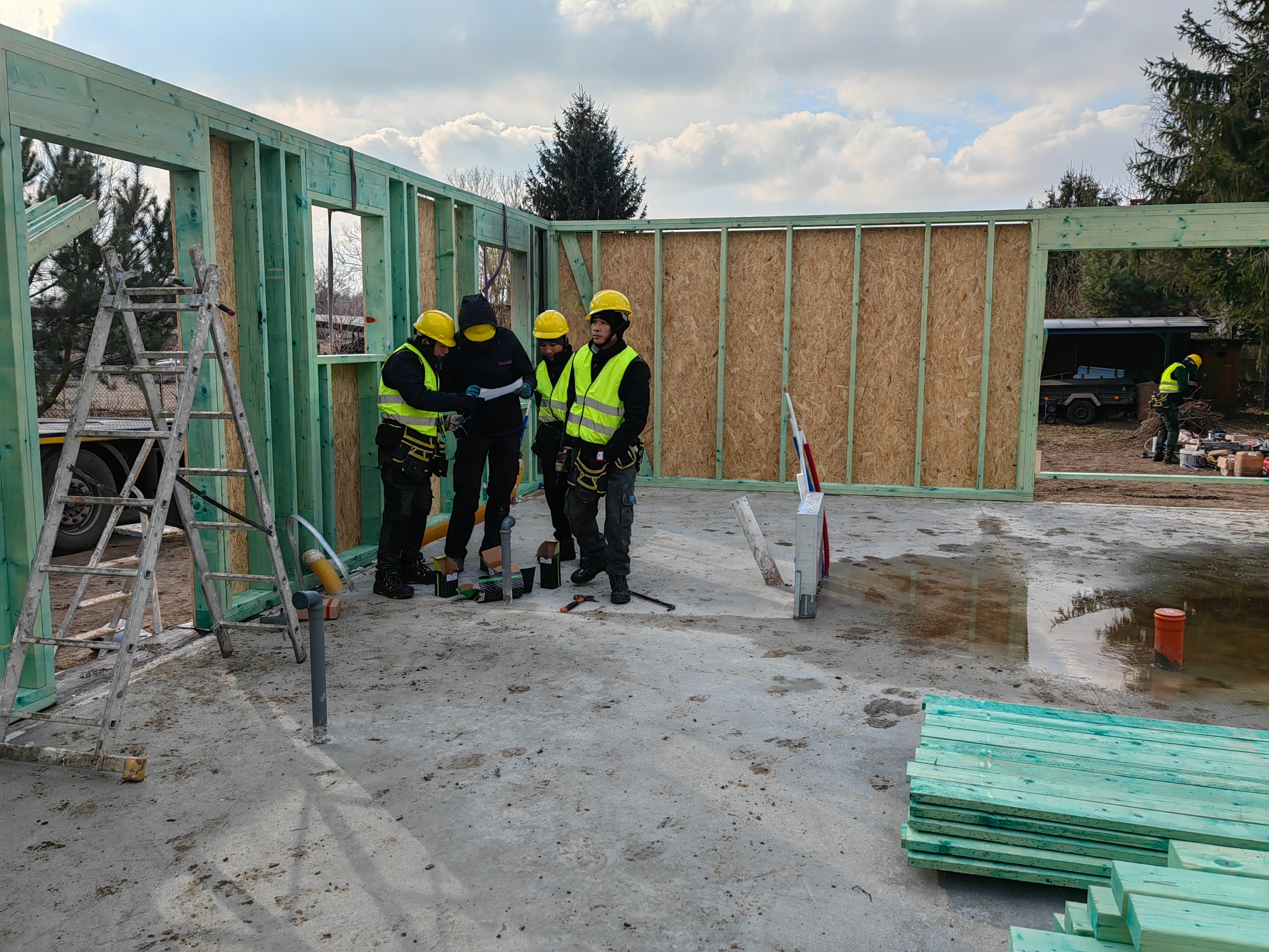 Ekipa budowlana w żółtych kaskach i kamizelkach, analizująca plany na budowie domu z drewnianą konstrukcją szkieletową, widoczna drabina i stos zielonych desek.