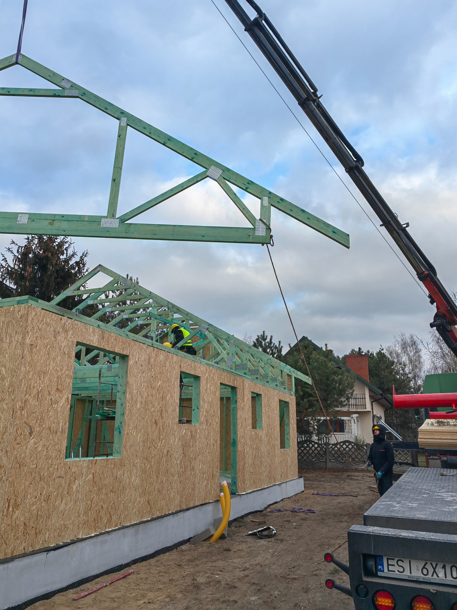 Montaż drewnianej konstrukcji dachu na budynku z płyt OSB, widoczny dźwig i pracownicy w kaskach na tle pochmurnego nieba.