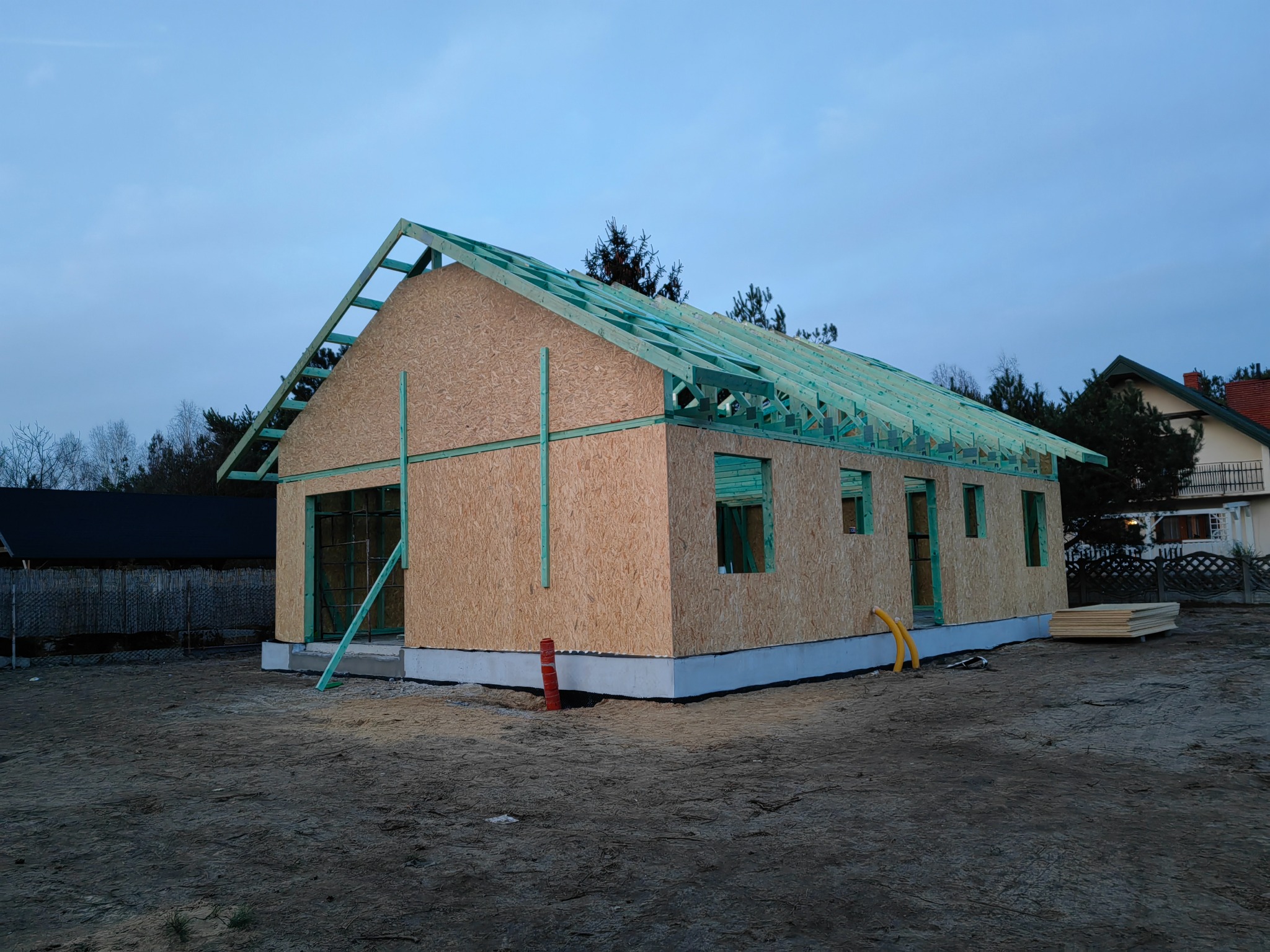 Surowa konstrukcja domu z płyt OSB i zieloną więźbą dachową na fundamencie, widok z zewnątrz w pochmurny dzień.