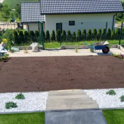 Przygotowany teren pod trawnik, obrzeża z białego kamienia i nasadzeniami, w tle dom z ciemnym dachem i zielone tuje wzdłuż ogrodzenia.