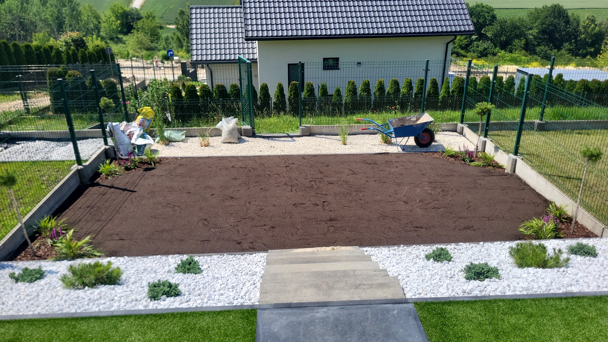 Przygotowany teren pod trawnik, obrzeża z białego kamienia i nasadzeniami, w tle dom z ciemnym dachem i zielone tuje wzdłuż ogrodzenia.