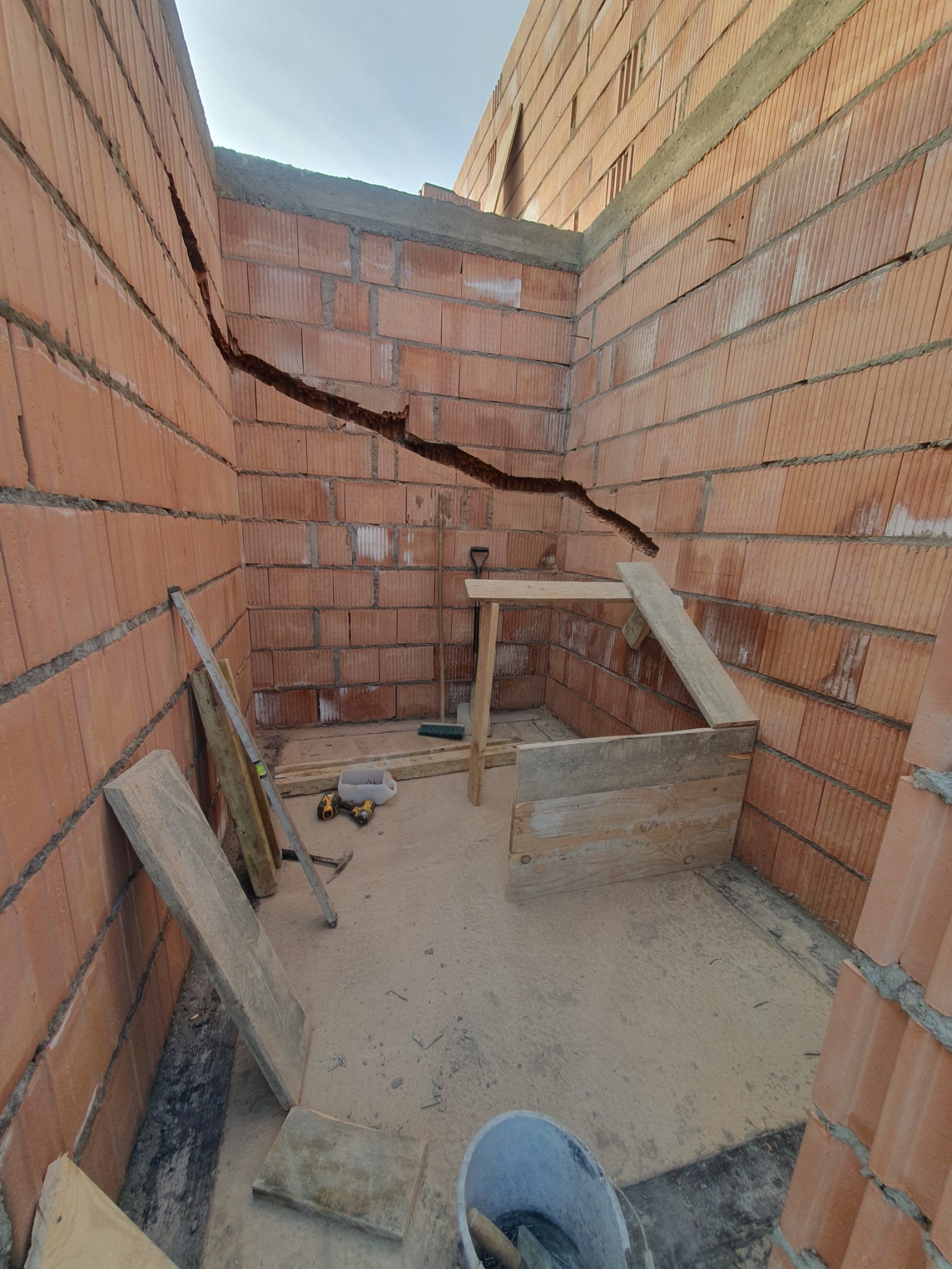 Wnętrze budynku w trakcie wznoszenia ścian z czerwonej cegły, widoczna duża pozioma szczelina w murze, drewniane stemple i narzędzia budowlane.