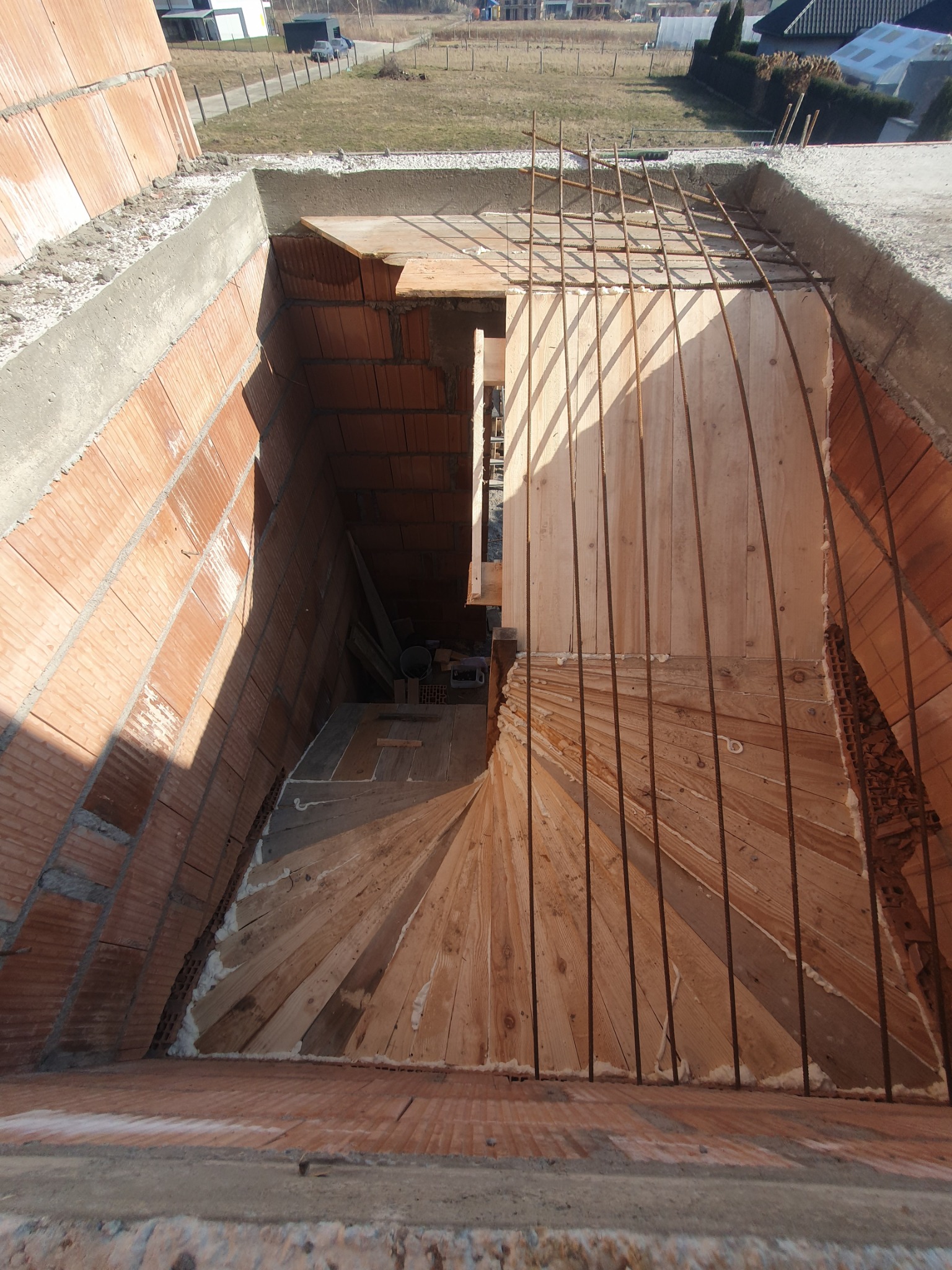 Wnętrze budynku w trakcie budowy, widoczne murowane ściany z pustaków ceramicznych, drewniane szalunki schodów spiralnych i zbrojenie stalowe, w tle widok na okolicę.