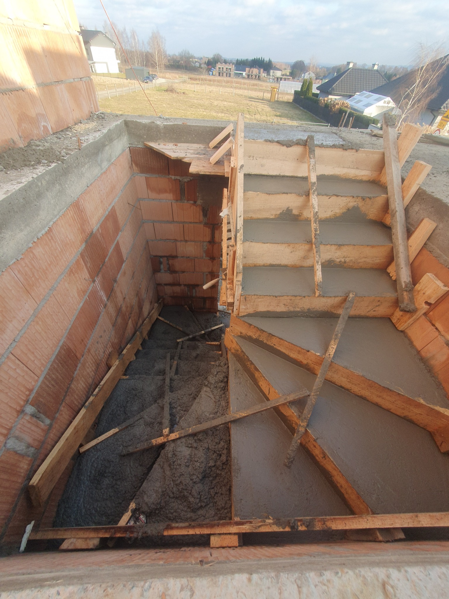 Wykonywanie schodów z betonu wewnątrz murowanej konstrukcji, widoczne drewniane szalunki i świeży beton, w tle zabudowa jednorodzinna.