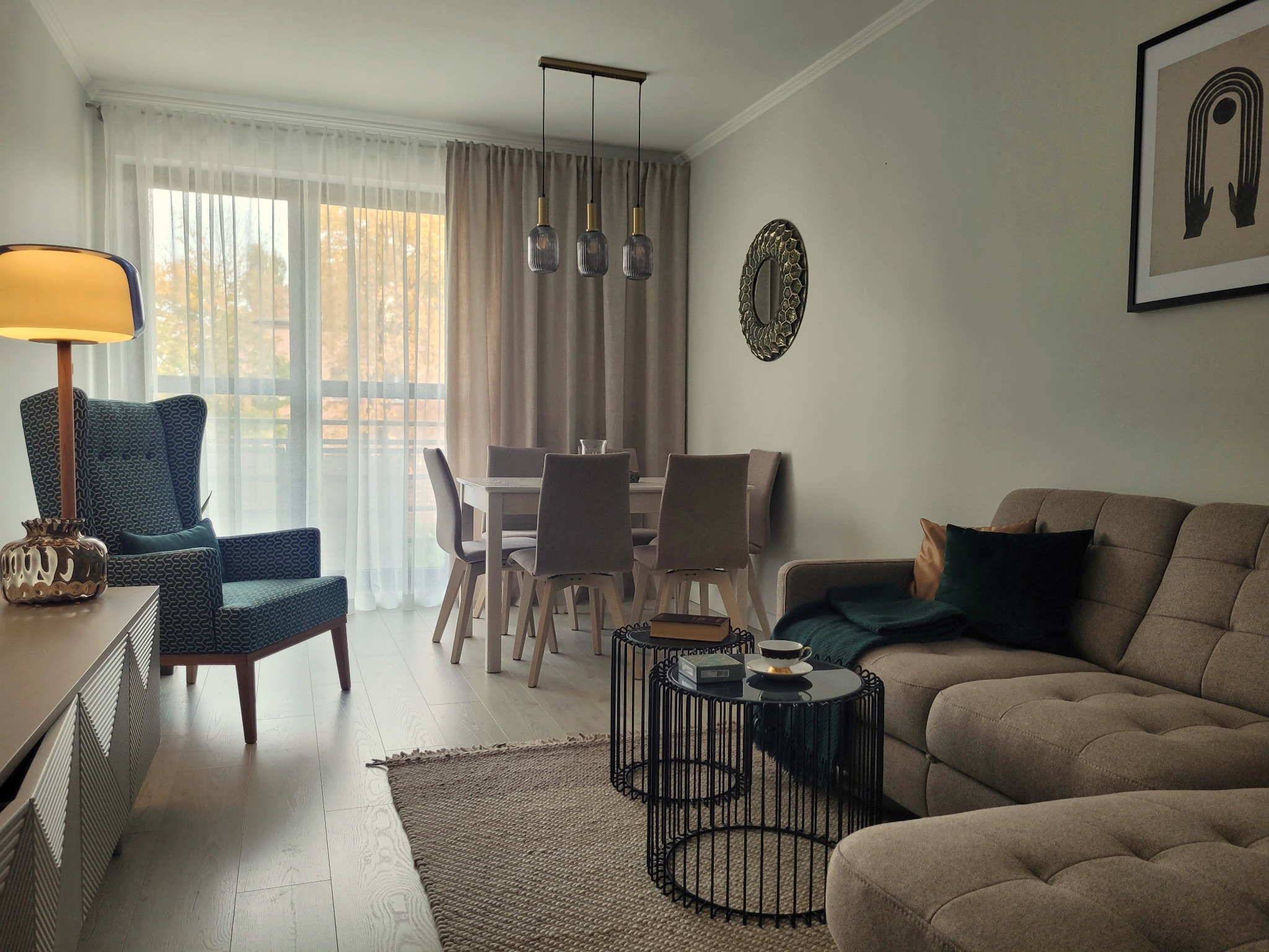 Jasne wnętrze salonu z jadalnią: szary narożnik, fotel w geometryczny wzór, okrągłe stoliki kawowe, stół z krzesłami pod trzypunktową lampą wiszącą, dywan z grubym splotem.