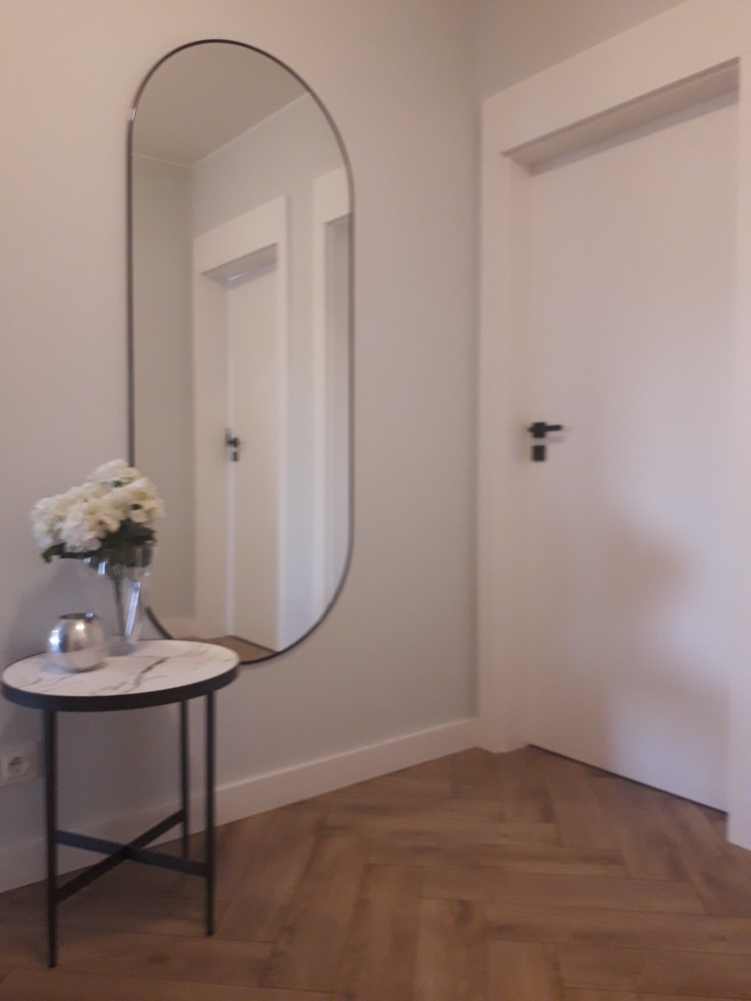 Nowoczesny przedpokój z parkietem ułożonym w jodełkę, owalnym lustrem z czarną ramą, stolikiem z marmurowym blatem i bukietem białych kwiatów, białe drzwi z czarną klamką.