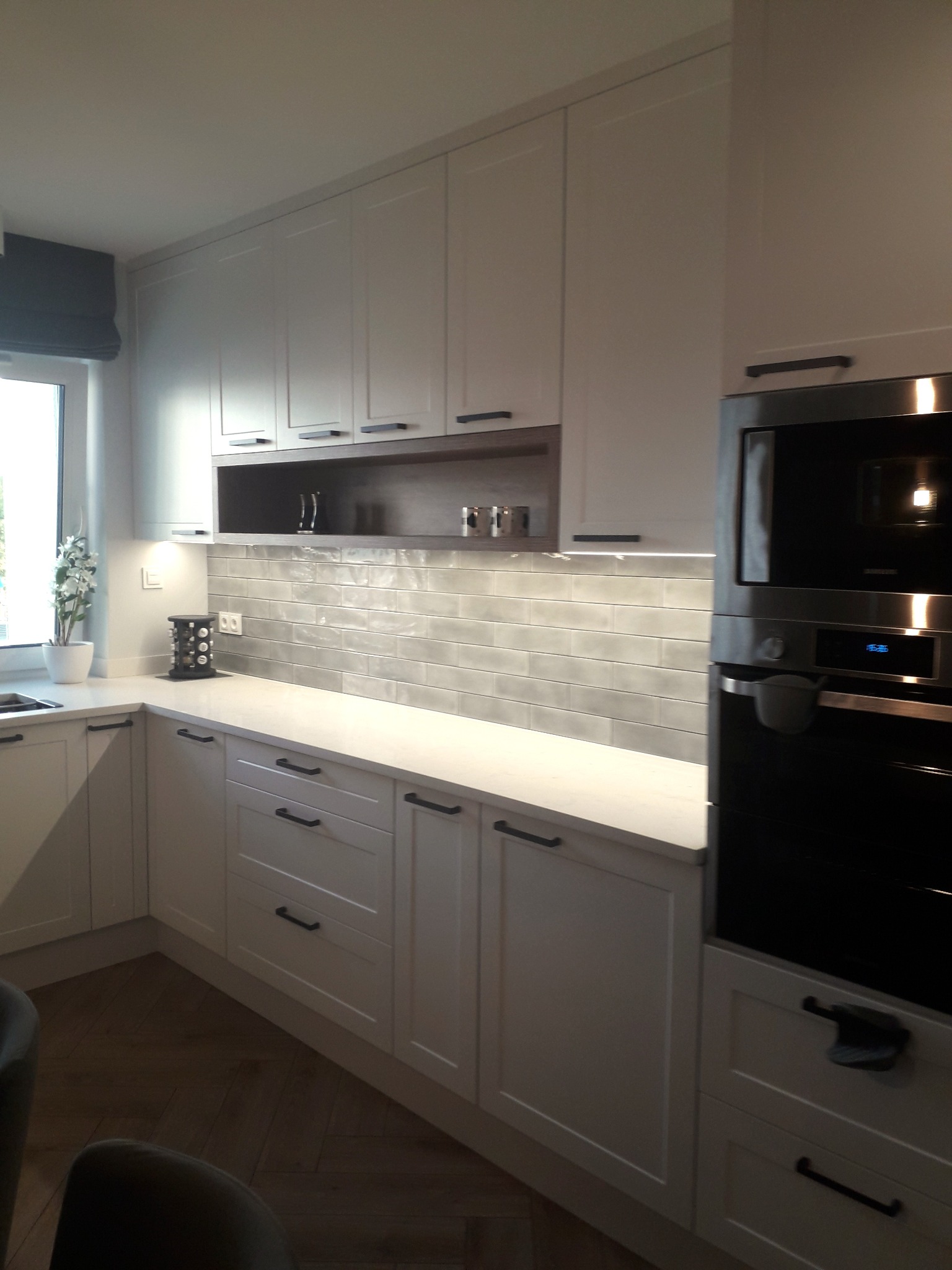 Jasna kuchnia z białymi szafkami, szarymi płytkami na ścianie i drewnianą podłogą ułożoną w jodełkę, oświetlona listwą LED pod szafkami wiszącymi.