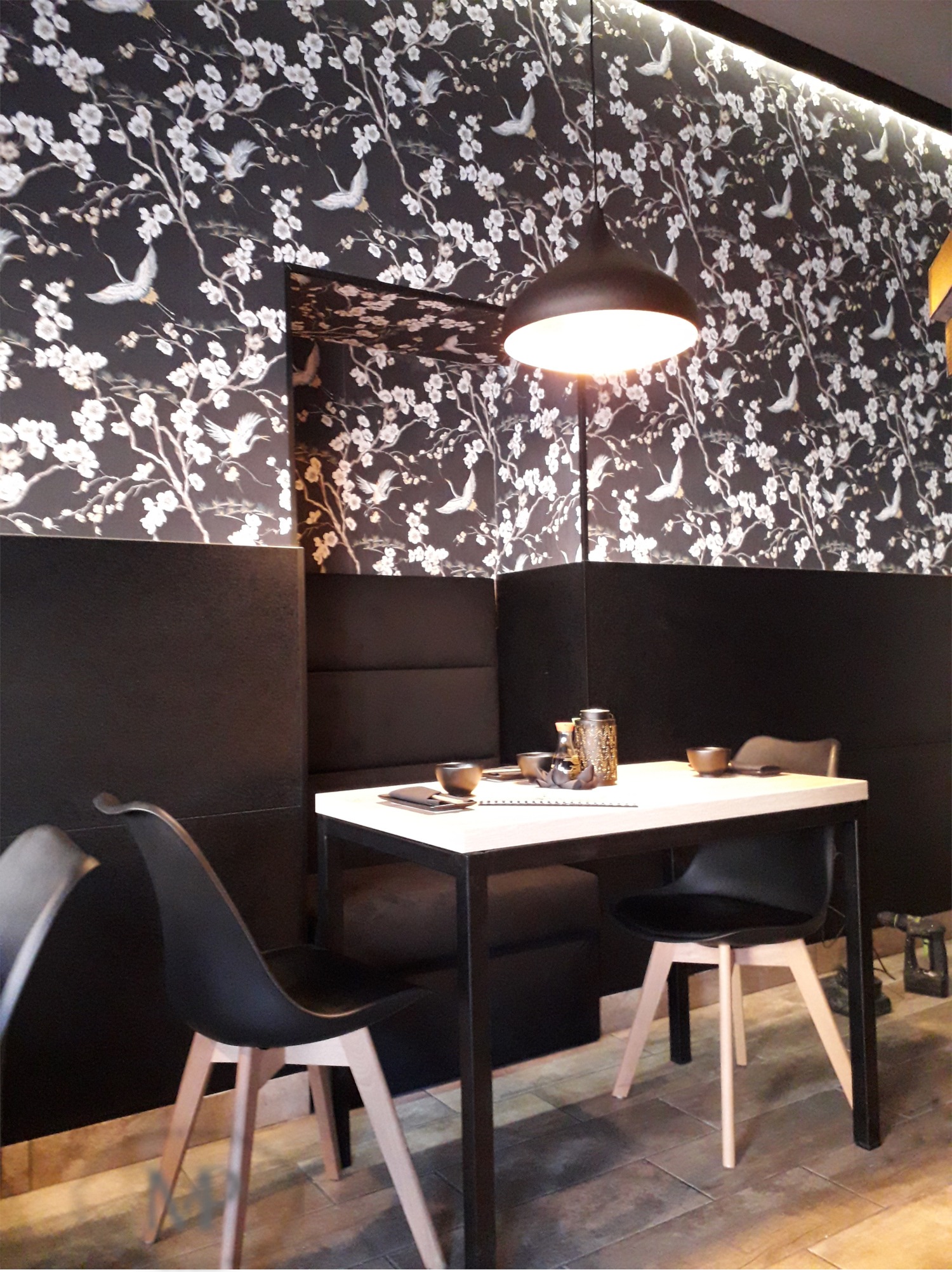 Stylowa aranżacja wnętrza restauracji w stylu orientalnym: tapeta z motywem kwitnących gałęzi i ptaków, czarne panele ścienne, stół z zastawą i nowoczesne krzesła z drewnianymi nogami.