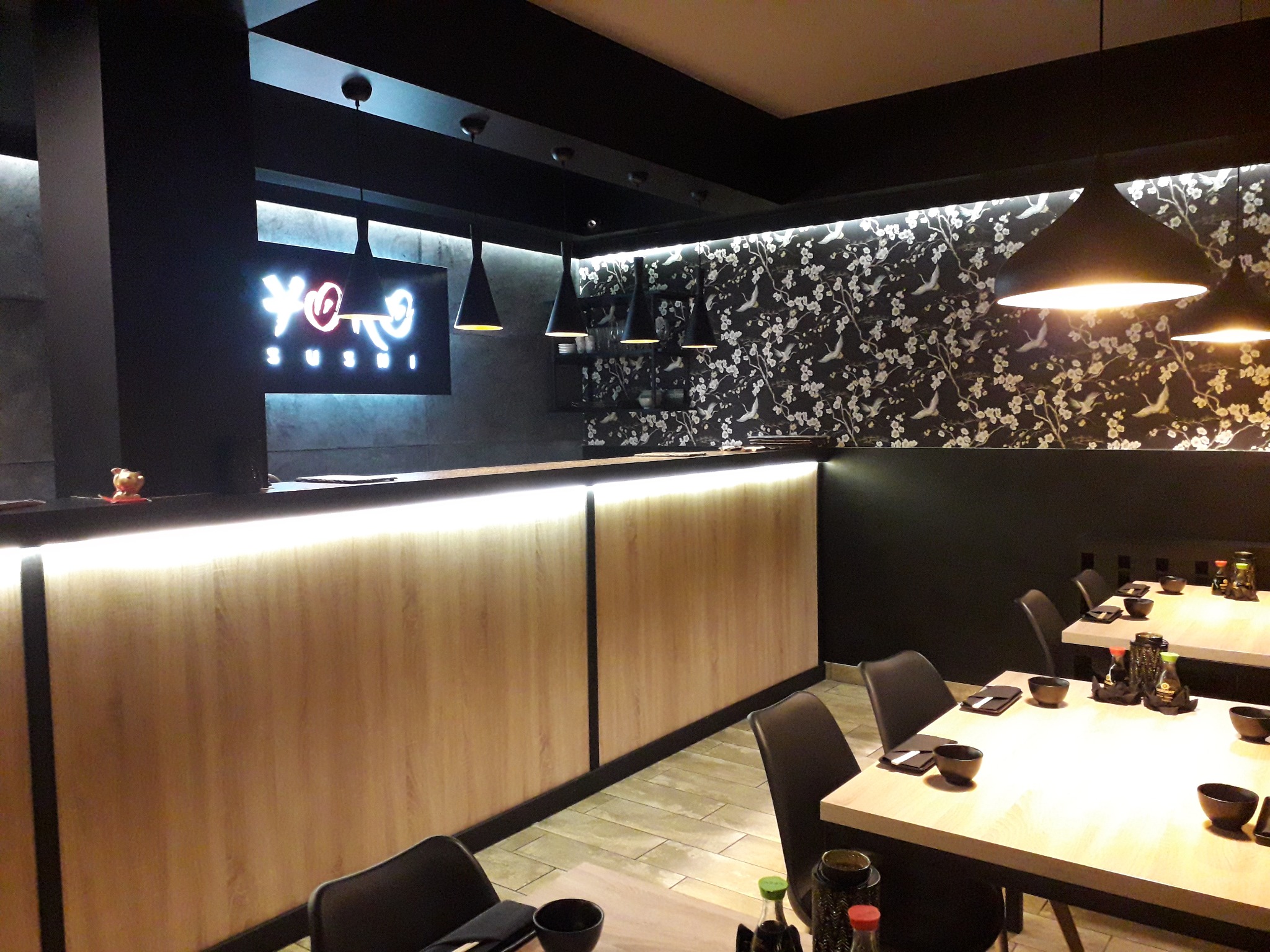 Wnętrze restauracji sushi z drewnianym barem podświetlonym ledami, czarne lampy stożkowe nad stołami i tapeta w motywy kwiatowe i ptasie.