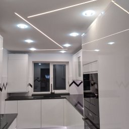 Krzysztof Sitniewski - Nowoczesna kuchnia w bieli z czarnym blatem i podłogą, oświetlenie sufitowe LED w geometryczne wzory, okno z czarną ramą i minimalistyczną armaturą.