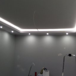 Krzysztof Sitniewski - Szare wnętrze po odświeżeniu z podwieszanym sufitem z oświetleniem LED i punktowym, widoczna drabina malarska i kabel zwisający z sufitu.