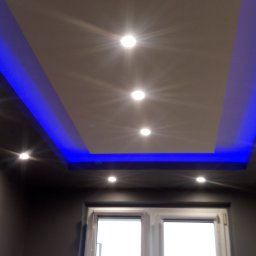 Krzysztof Sitniewski - Podwieszany sufit z oświetleniem LED w kolorze niebieskim i centralnie umieszczonymi punktowymi lampami, widok z dołu na tle okna.