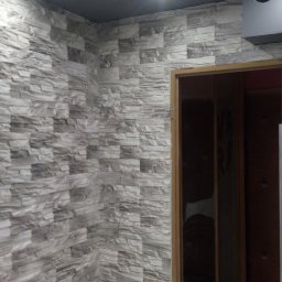 Krzysztof Sitniewski - Ściana wykończona panelami imitującymi kamień w odcieniach szarości, oświetlona punktowymi lampami sufitowymi, widok na ościeżnicę drzwiową z fragmentem ciemnej podłogi w tle.